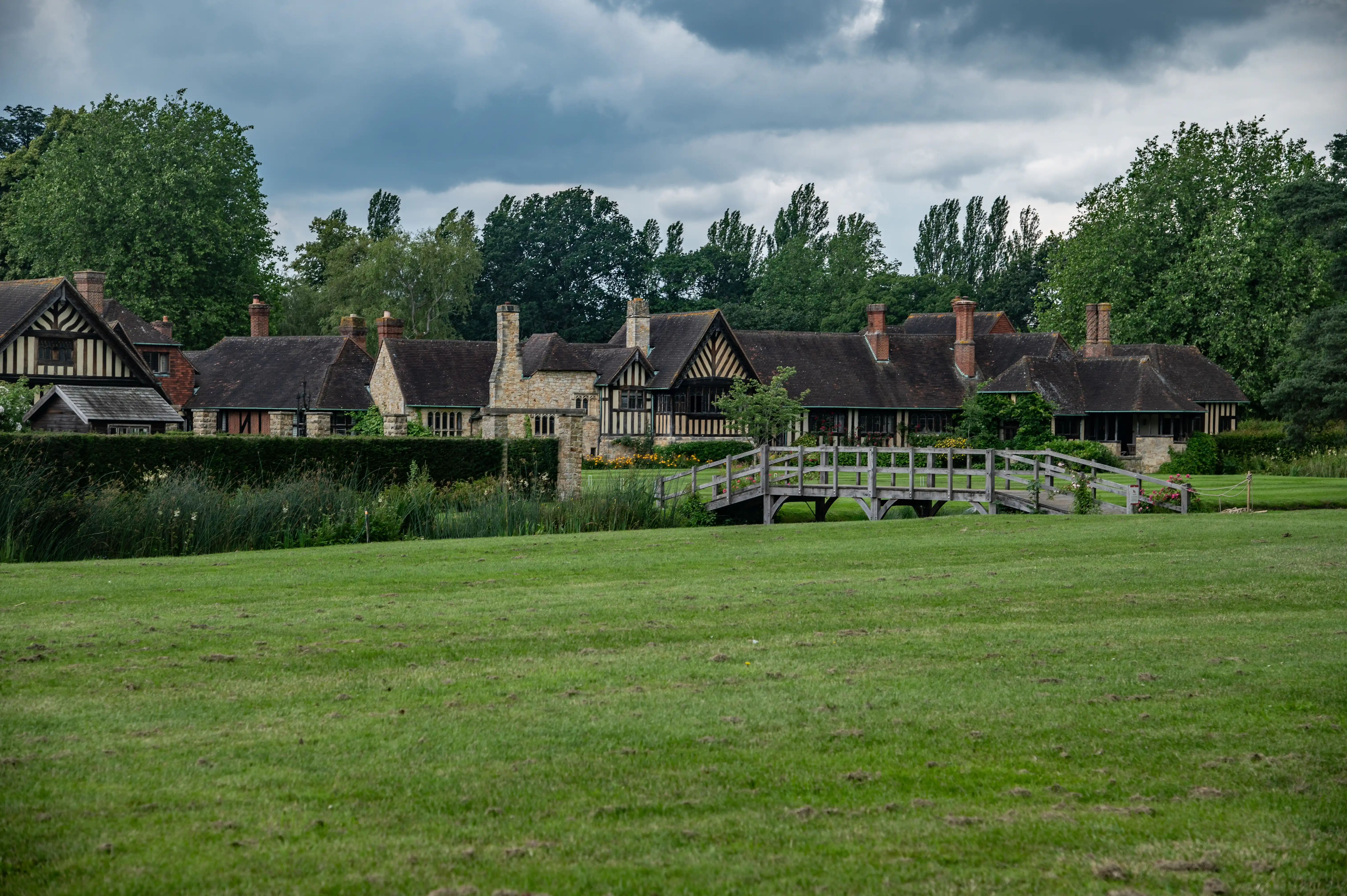 A traditional English country house with dark tiled roofs and exposed timber beams, viewed from a manicured green lawn. A quaint wooden bridge crosses a narrow body of water in front of the house, with mature trees forming a backdrop under a cloudy sky.