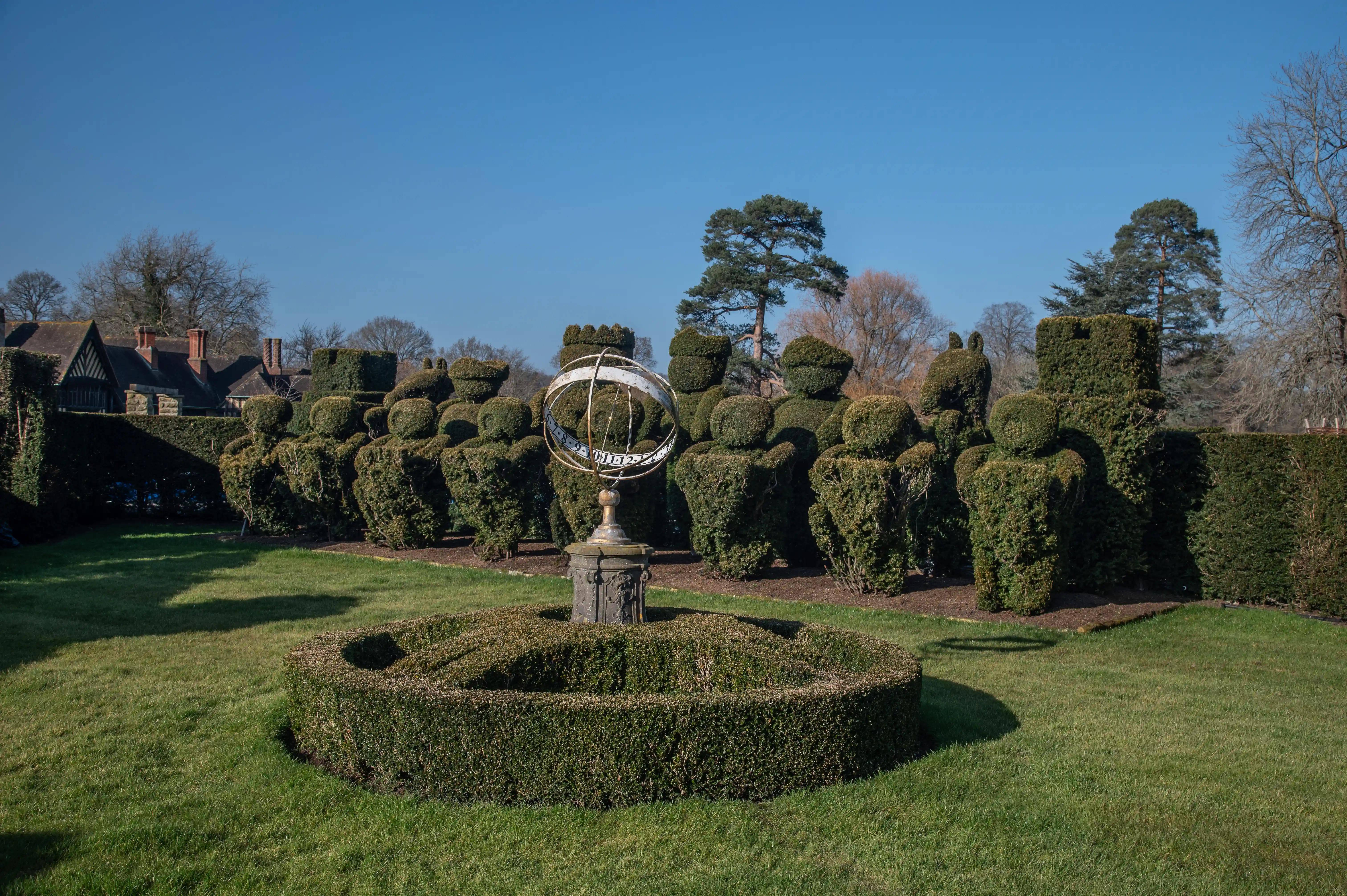 A clear, sunny day illuminates a formal garden featuring a row of meticulously sculpted topiary figures resembling people, standing behind a sundial situated in a circular, low-lying hedge. In the background, a traditional building with a pitched roof and chimneys can be seen, along with mature trees.