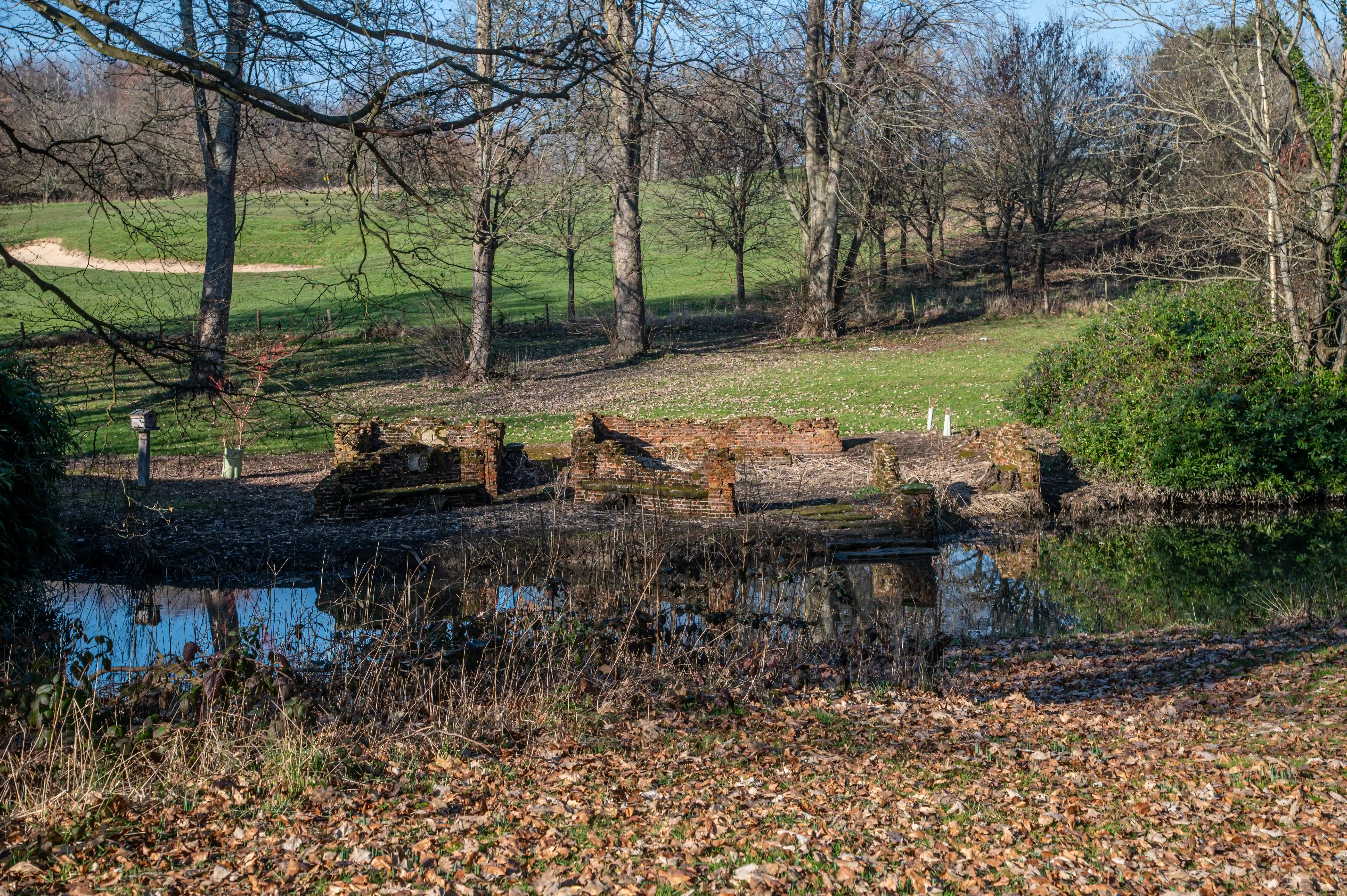 Old brick ruins reflected in a dark pond, surrounded by bare trees and a carpet of autumn leaves, with a green grassy hill in the background under a blue sky.