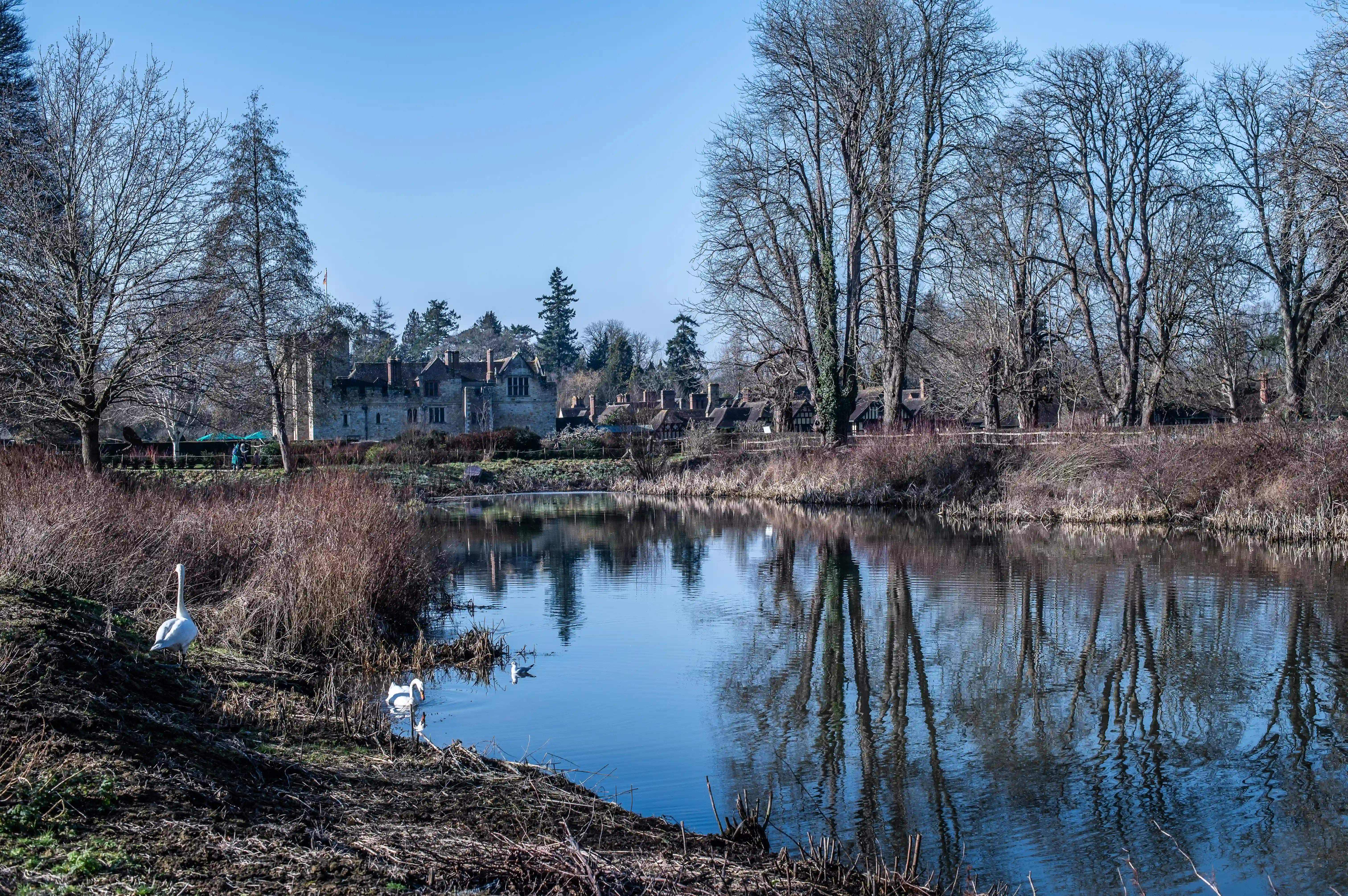 A low-angle shot of a quaint wooden and brick bridge arching over a still river. The bridge's reflection is visible in the water, surrounded by bare trees and under a bright blue sky.