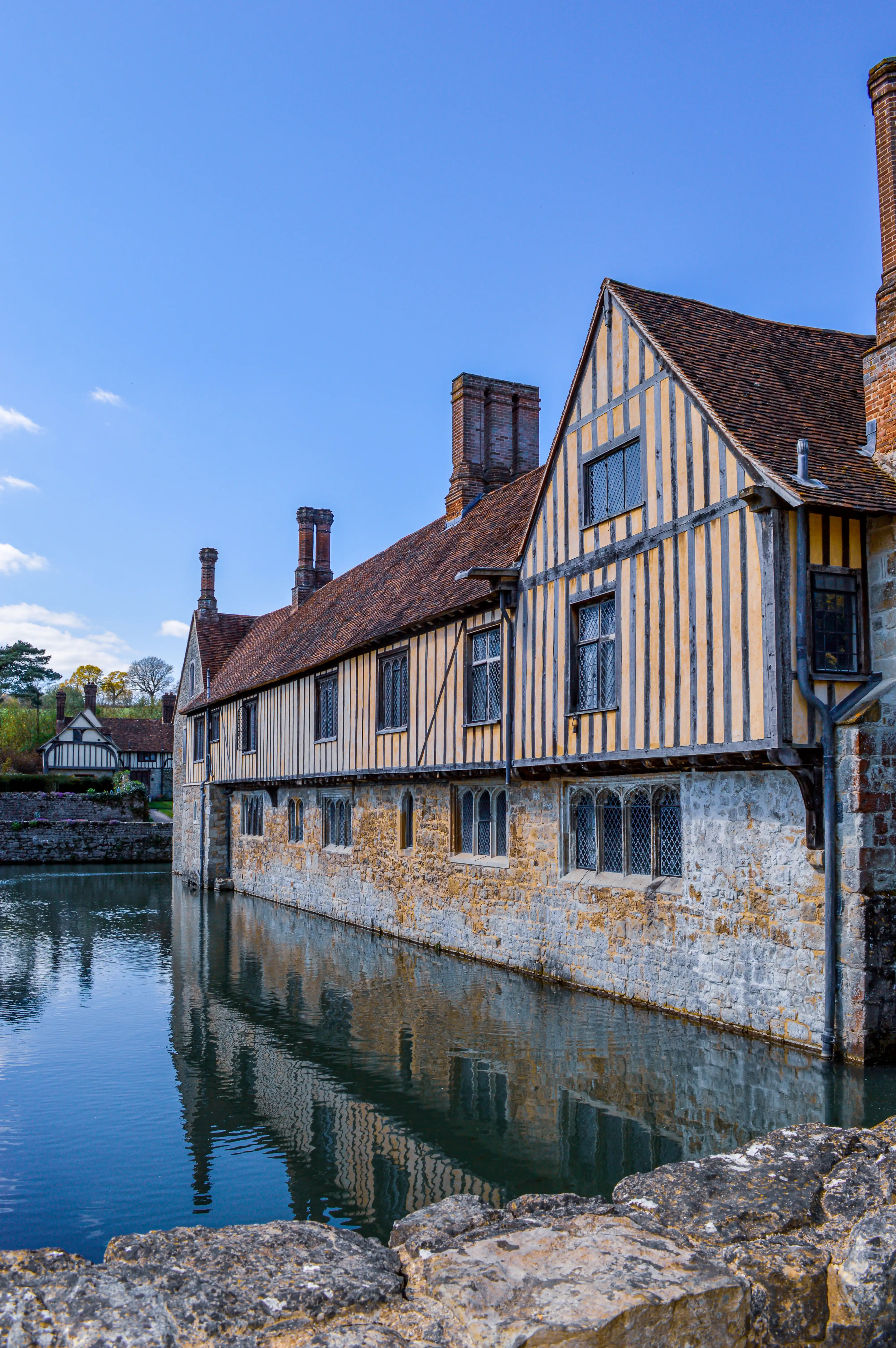 A historic Tudor building with a stone base and timber-framed upper storeys, reflected in a calm moat under a bright blue sky.