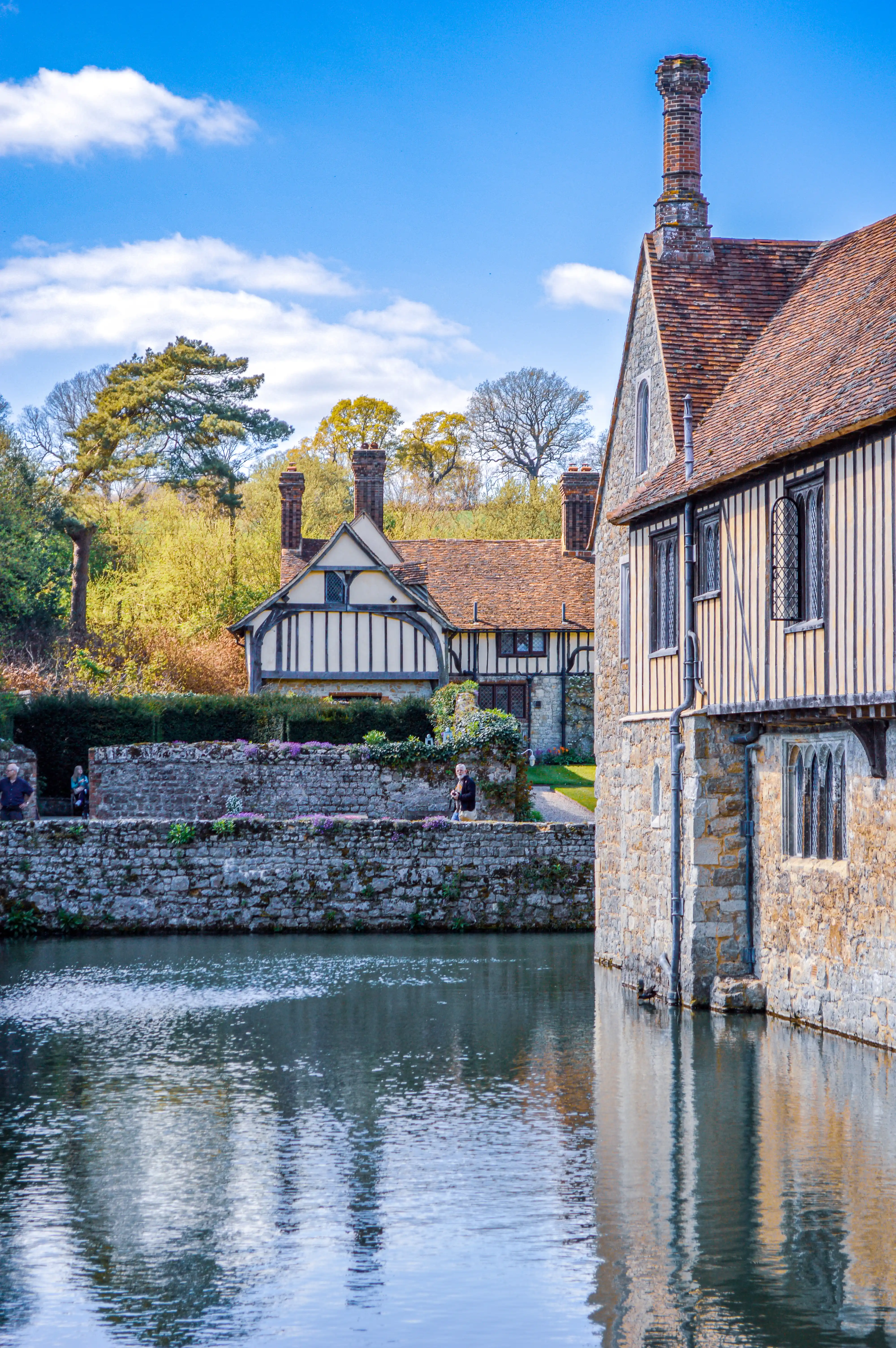 Historic timber-framed manor house with a stone moat wall, purple flowers, and reflections in the water under a blue sky.