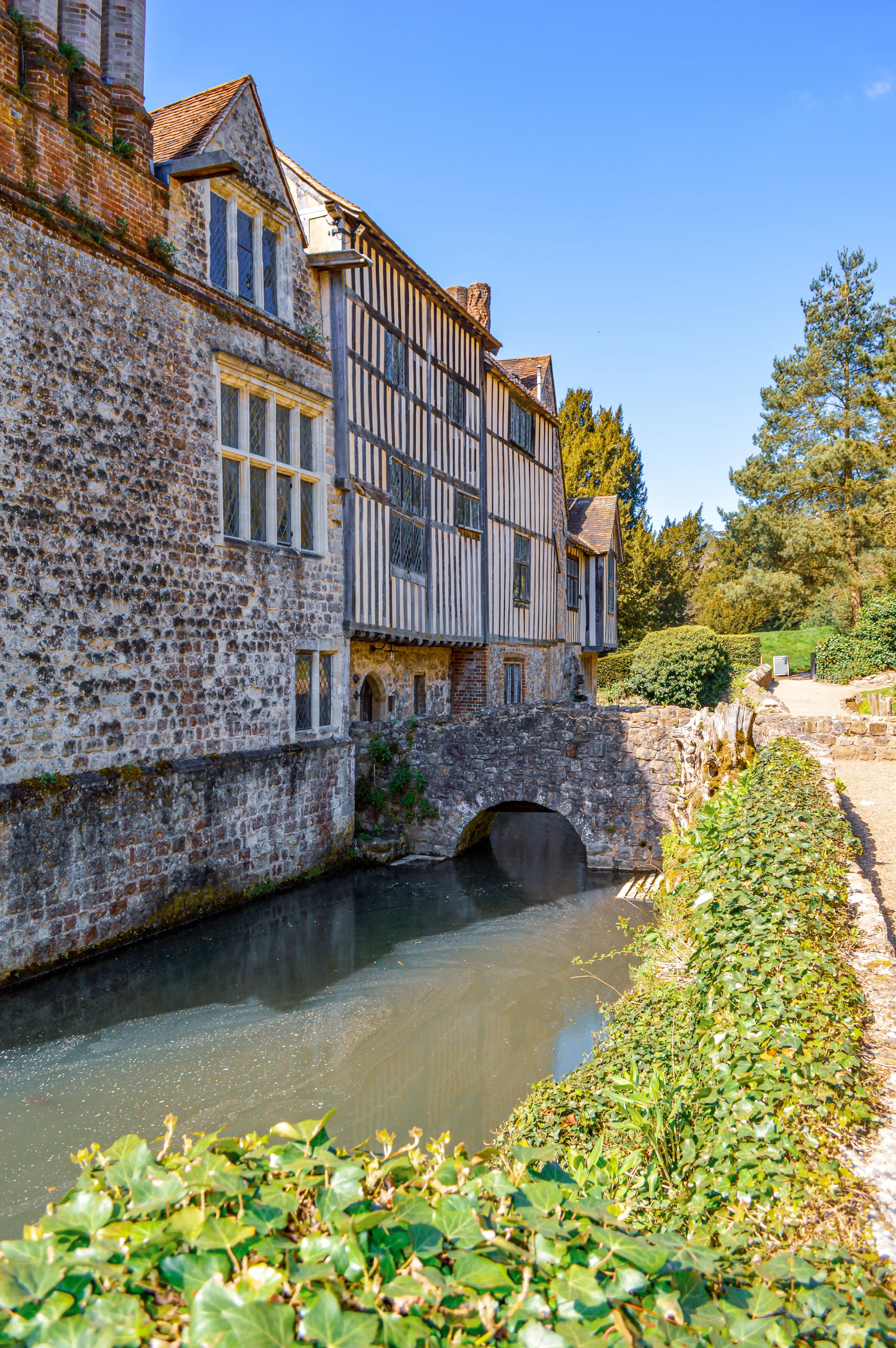 A half-timbered historic building with stone foundations stands next to a reflective moat, crossed by an ancient stone arched bridge. Green foliage and trees surround the scene under a bright blue sky.