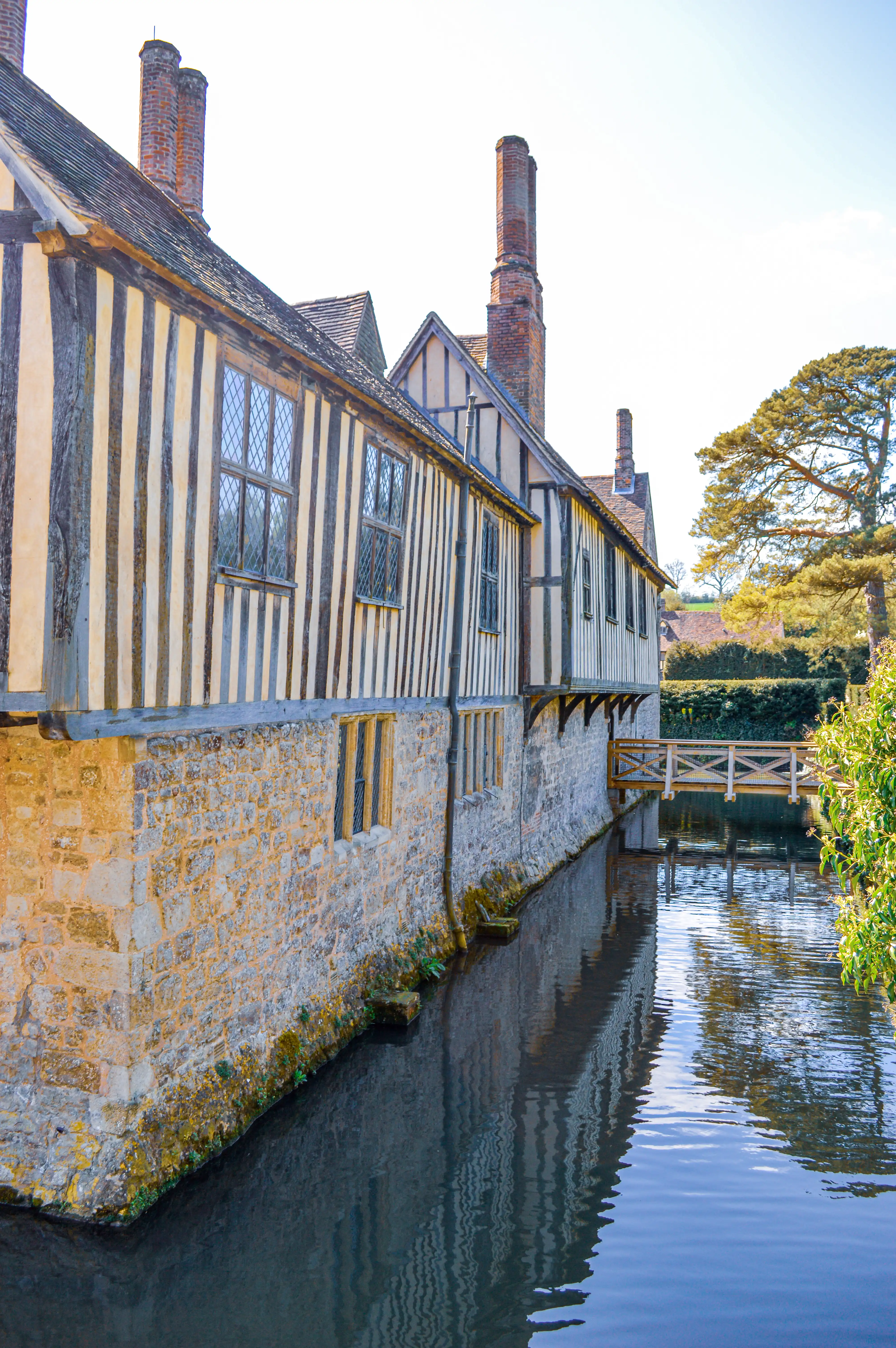 A long, two-storey timber-framed building with a stone lower level stands next to a dark, reflective body of water. A wooden bridge spans the water. Tall brick chimneys rise from the roof, and leaded glass windows are visible. Green foliage and a large tree are to the right of the building.