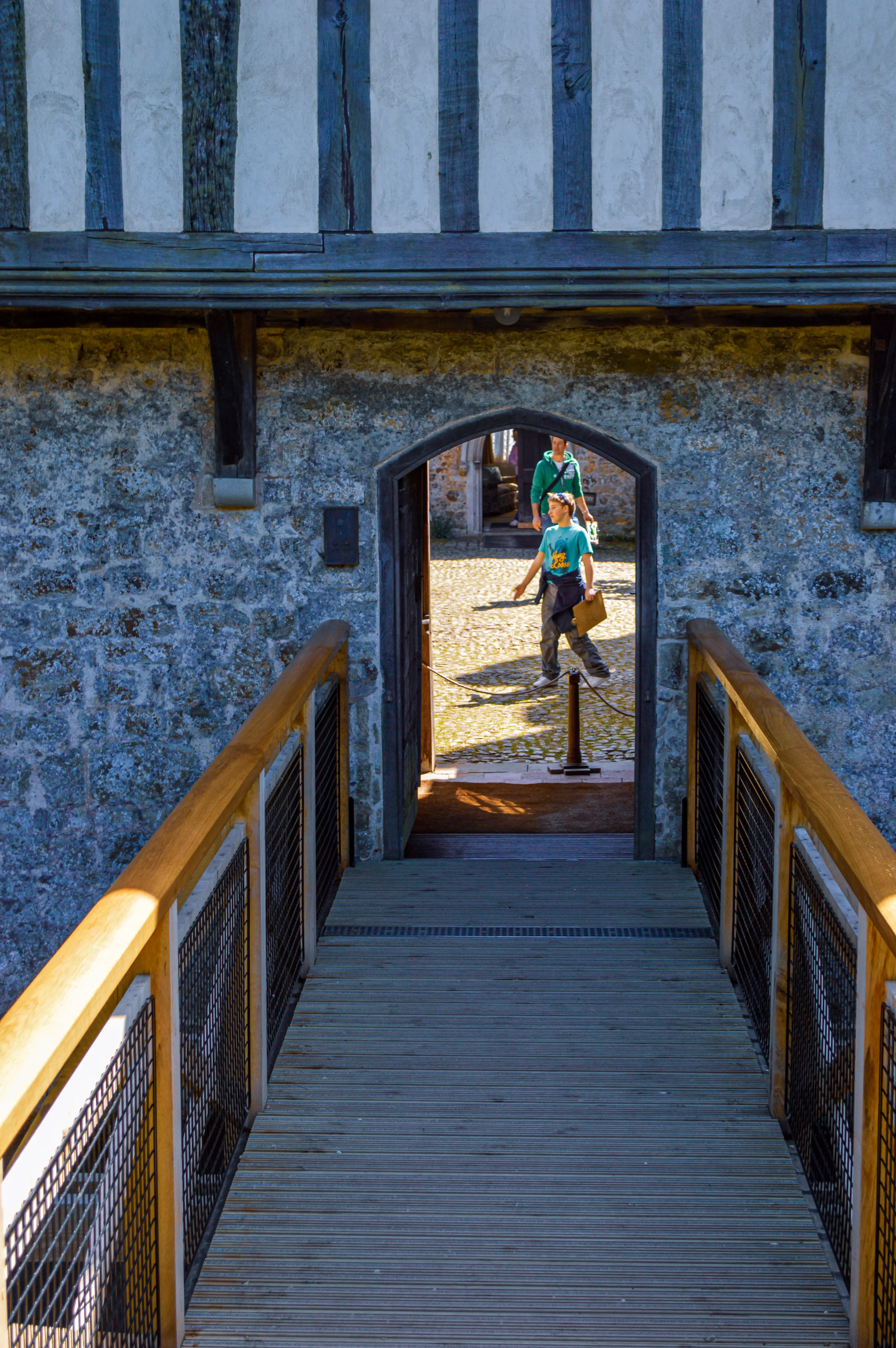 A person in a teal t-shirt and grey trousers walks through an arched stone doorway into a courtyard, with a wooden bridge leading up to the entrance.