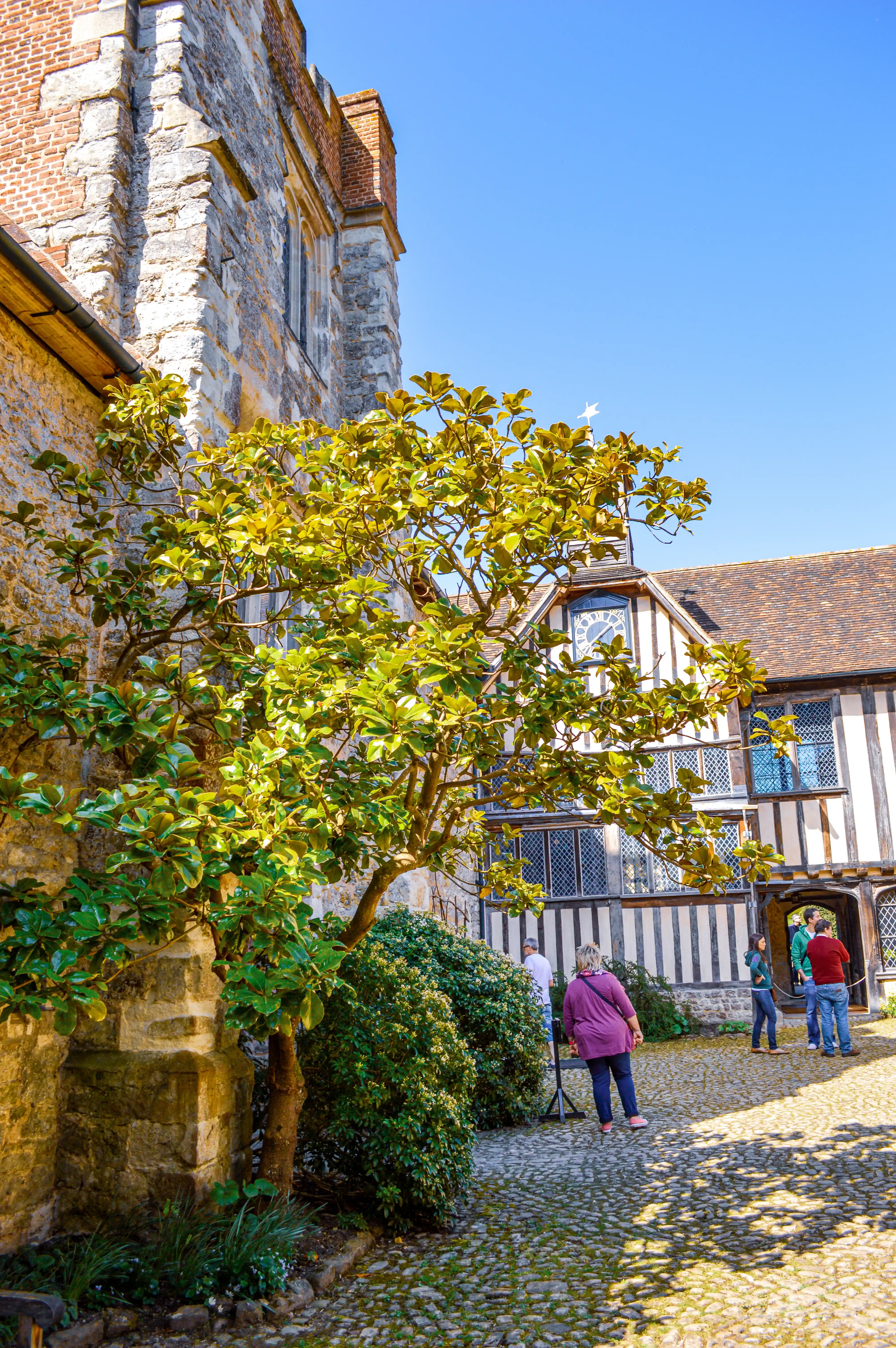 People walking in a courtyard of a historic timber-framed building under a clear blue sky.