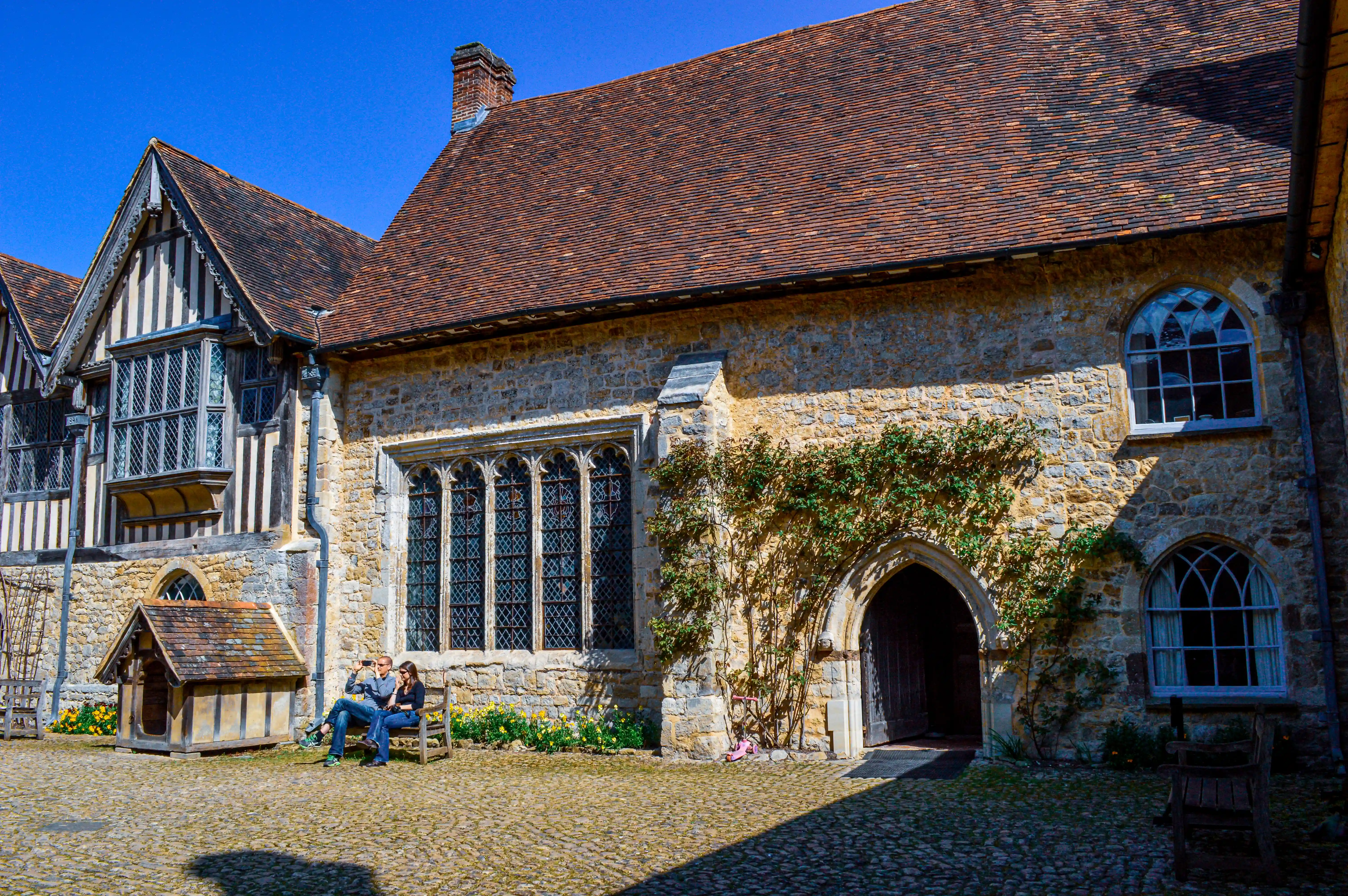 A couple sits on a wooden bench in a cobblestone courtyard in front of a historic stone building with a large arched doorway and leaded glass windows.