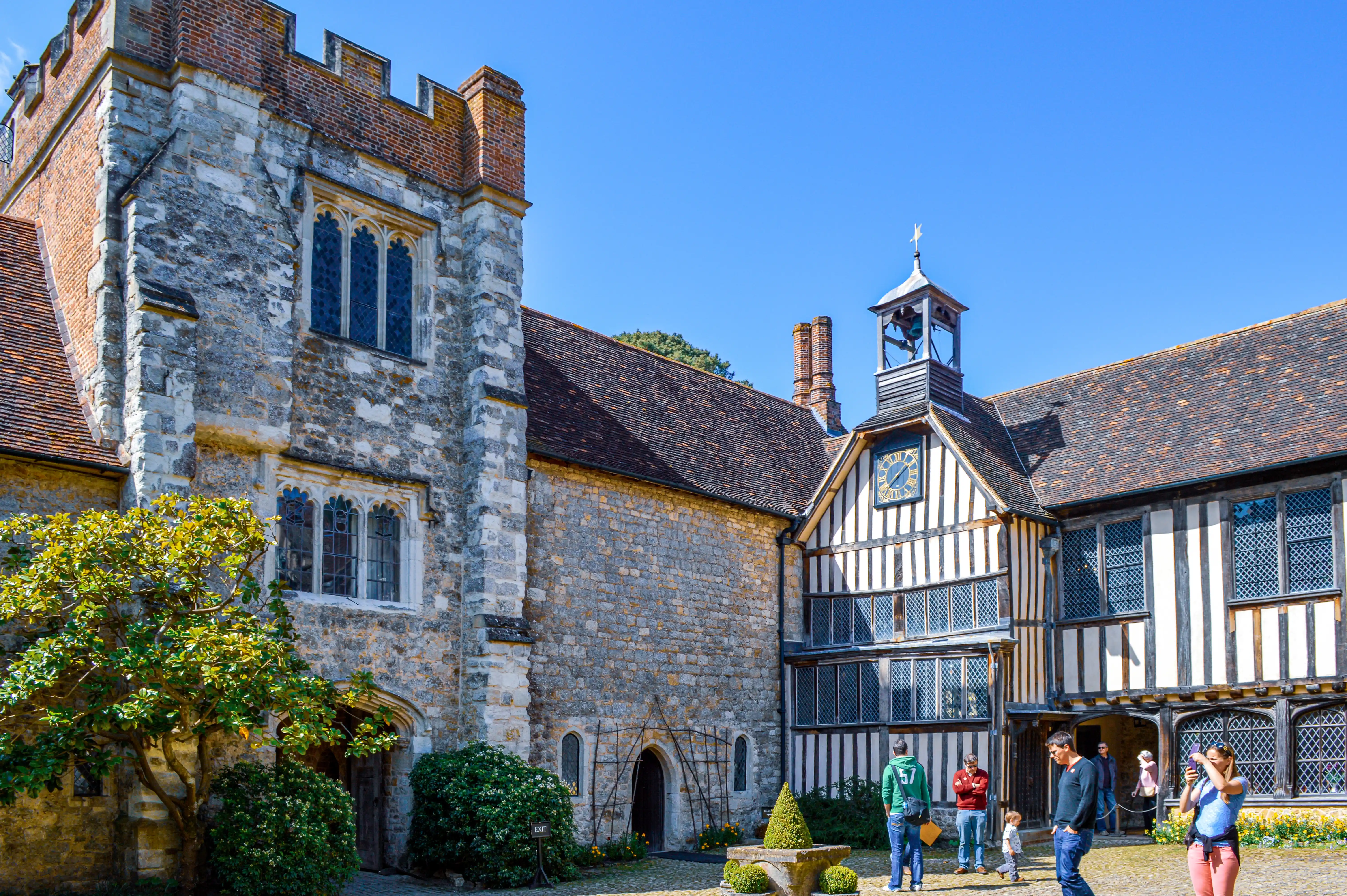 A courtyard of a historic building with a stone section on the left and a timber-framed section with a clock on the right. People are standing in the courtyard, and the sky is clear blue.