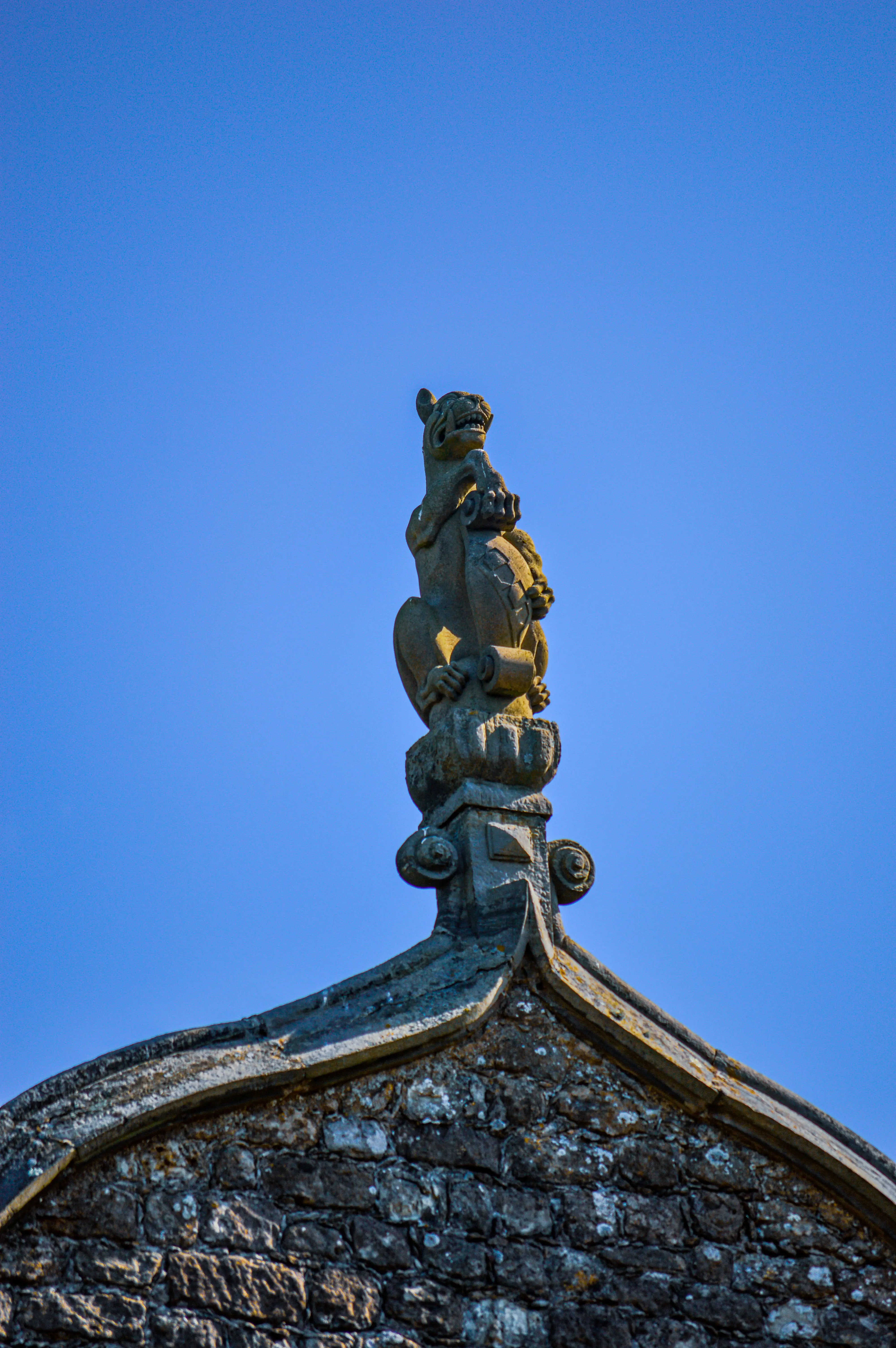 A stone gargoyle or griffin sculpture with its mouth open and paws resting on a shield, set against a vivid blue sky. The sculpture sits atop a decorative stone structure on the peak of a stone building.