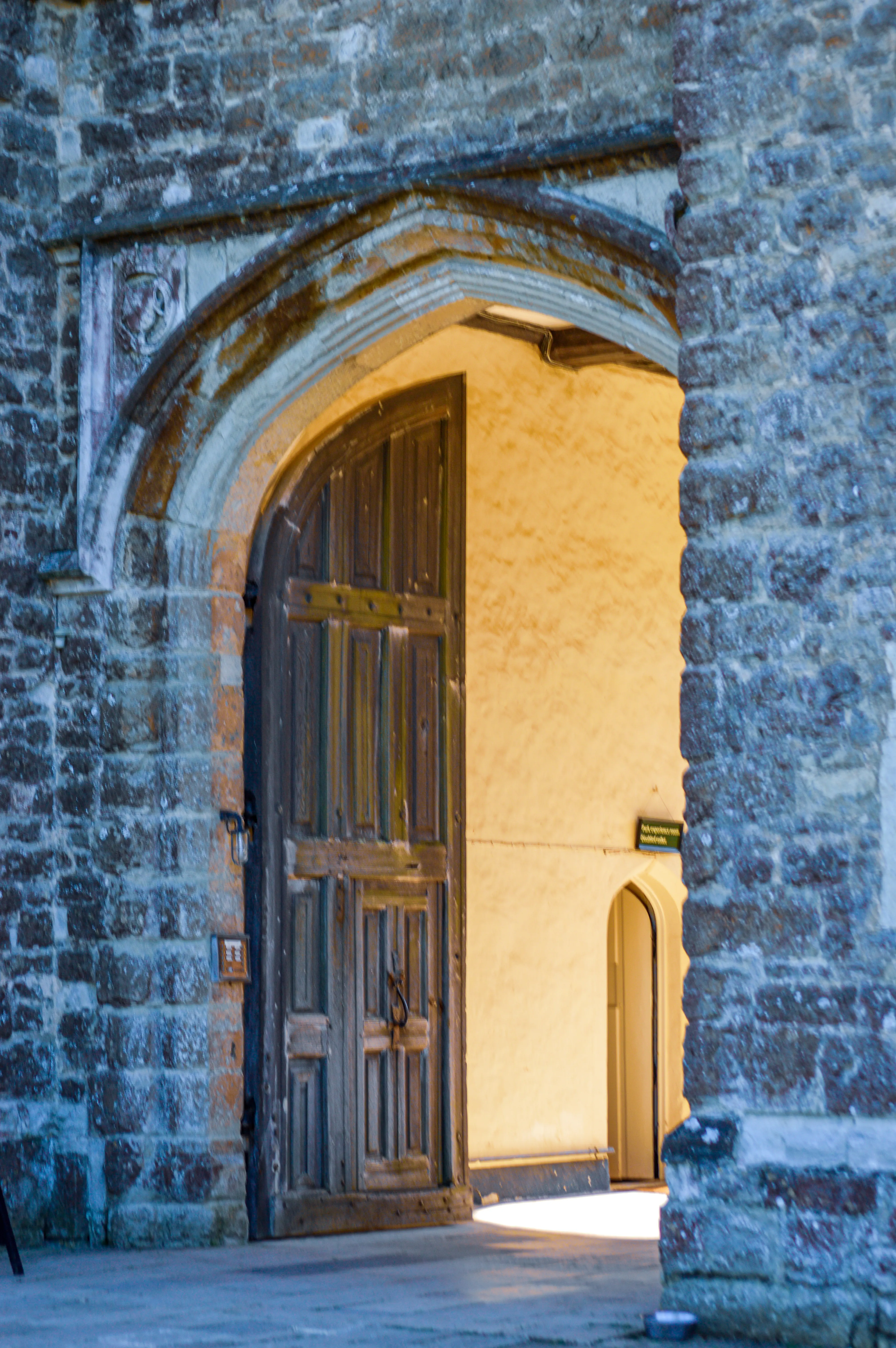 A stone archway frames a partially open, dark wooden door. The stonework is rough-hewn and ancient. Beyond the door, a brightly lit interior with another smaller, arched doorway is visible.