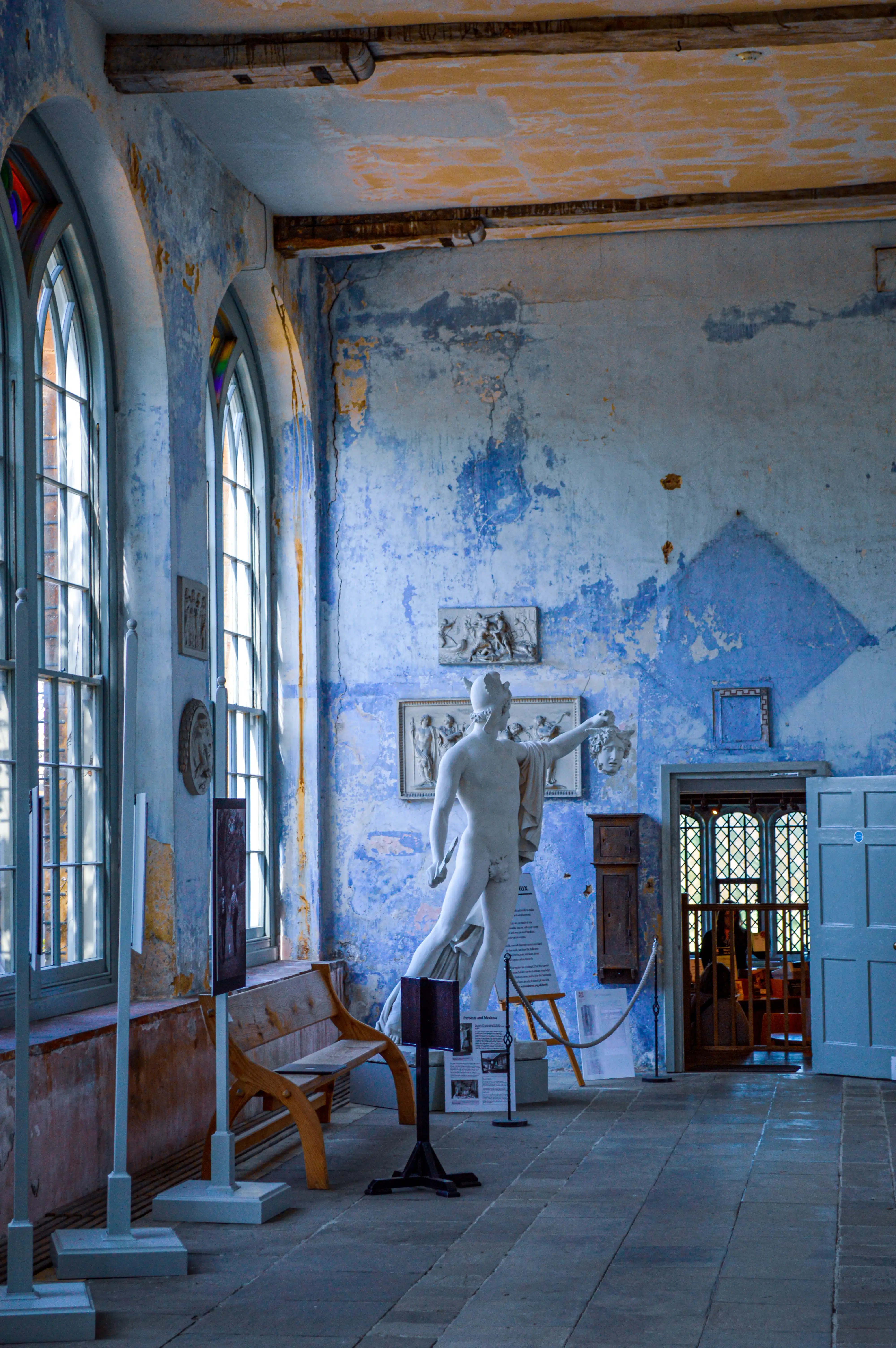 A white marble statue of a male figure holding a severed head, positioned in a room with distressed blue walls, arched windows, and wooden beams overhead.
