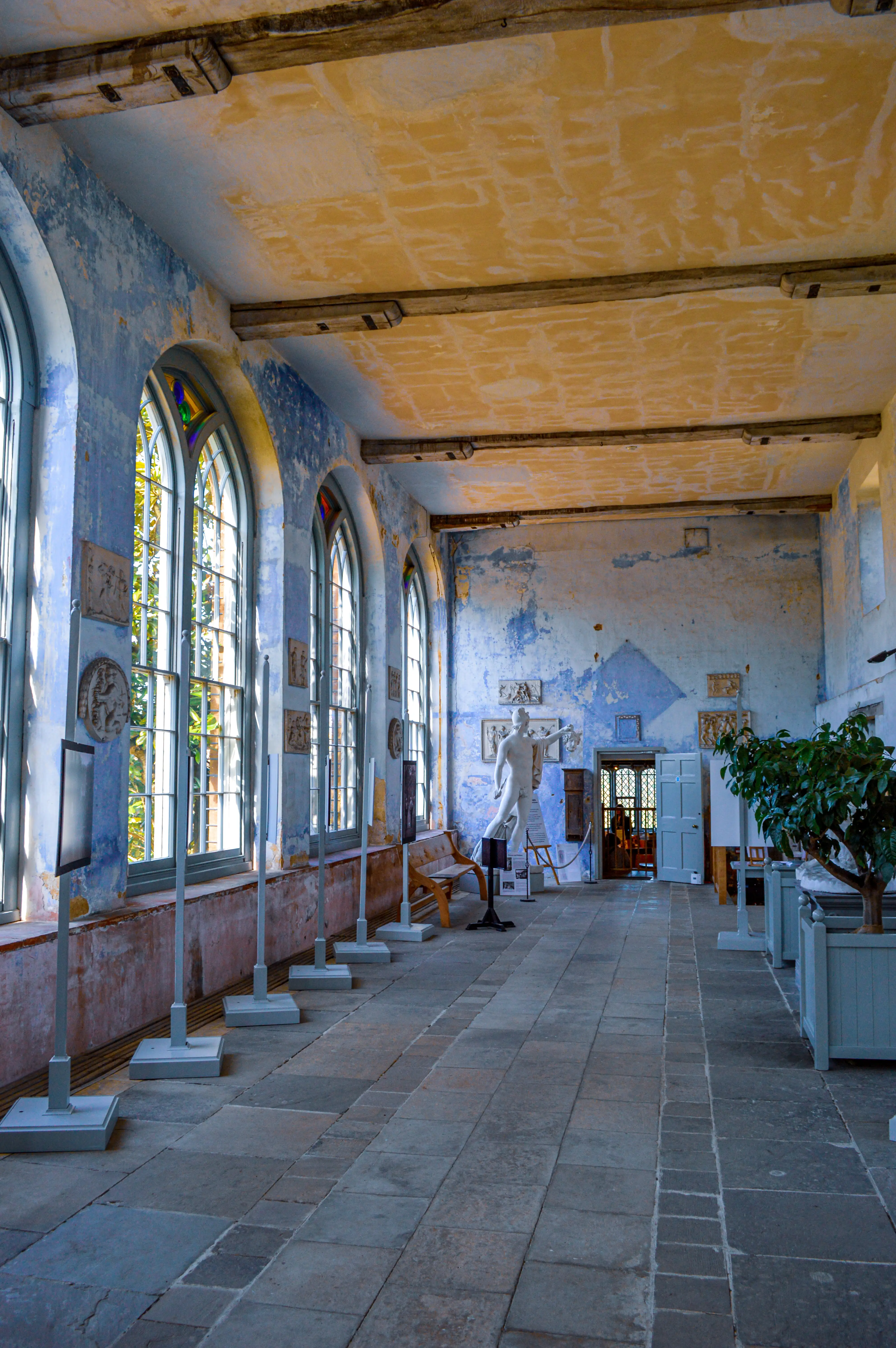 Interior of a historical gallery space with peeling blue walls, tall arched windows letting in sunlight, a classical white sculpture, and a stone floor.