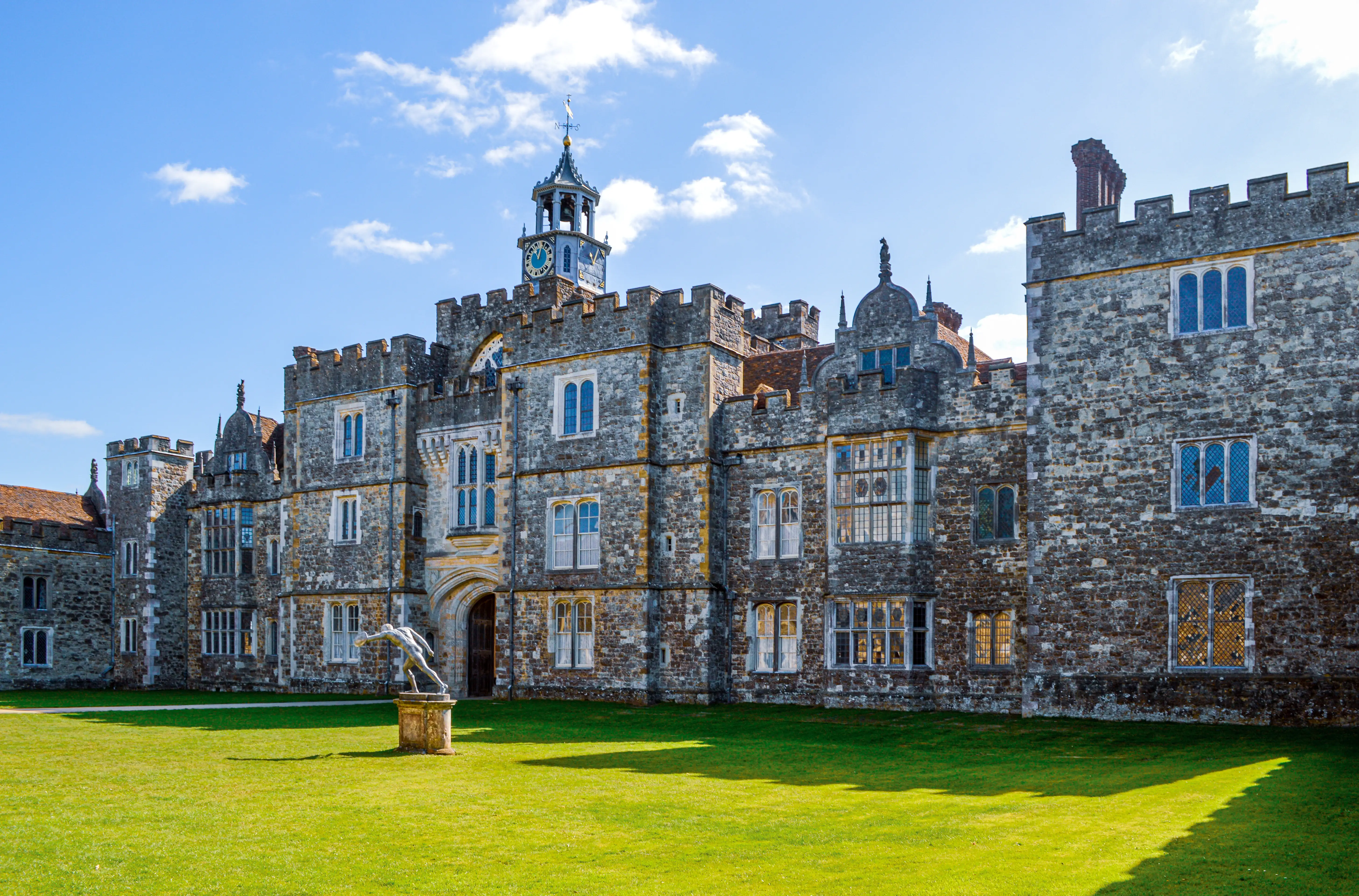 A majestic stone manor house with a clock tower and a statue of a man on a pedestal in front of it, set against a blue sky.