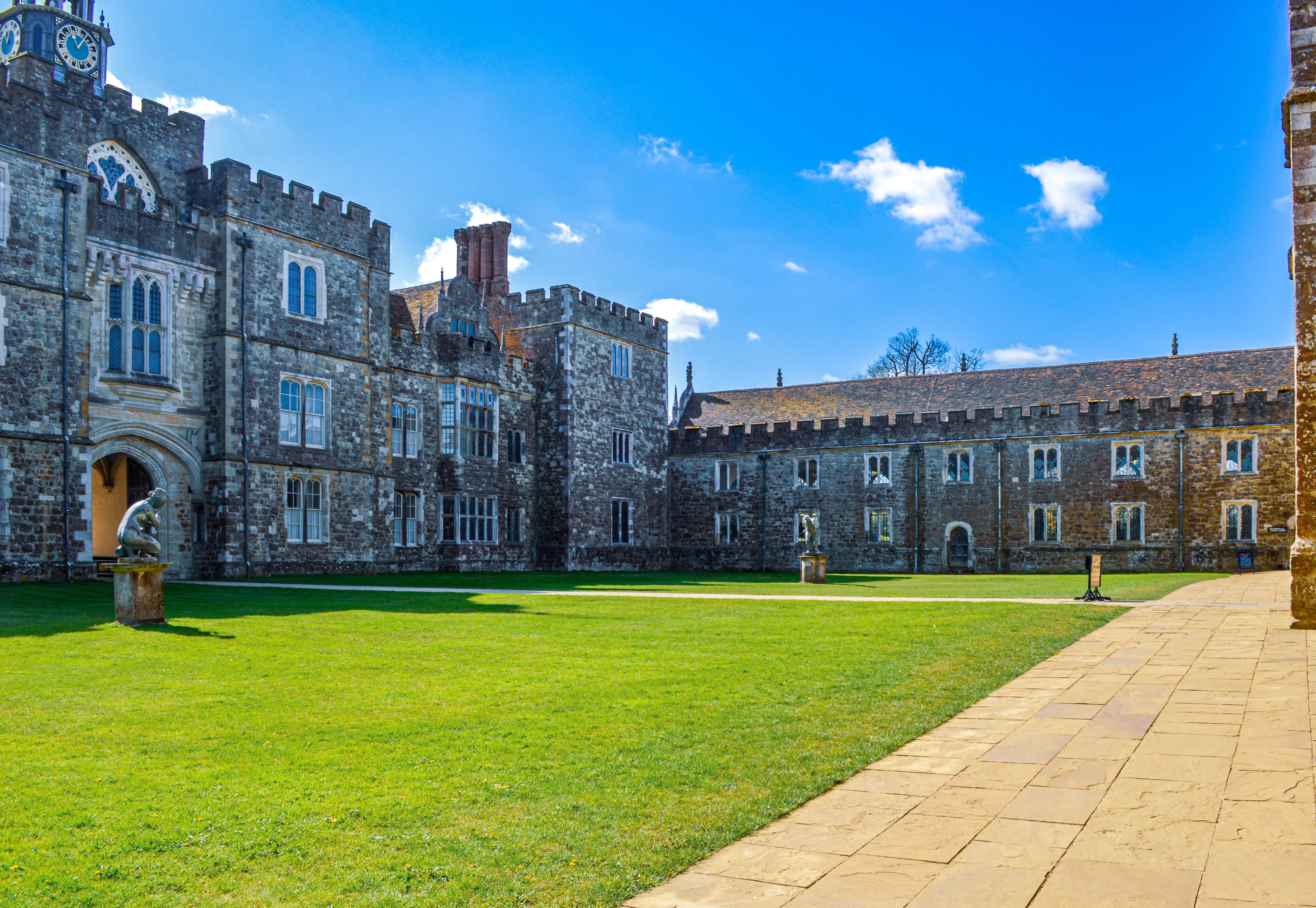 A wide shot of a grand, old stone building with a clock tower and crenellated battlements, viewed across a lush green lawn and a paved pathway.