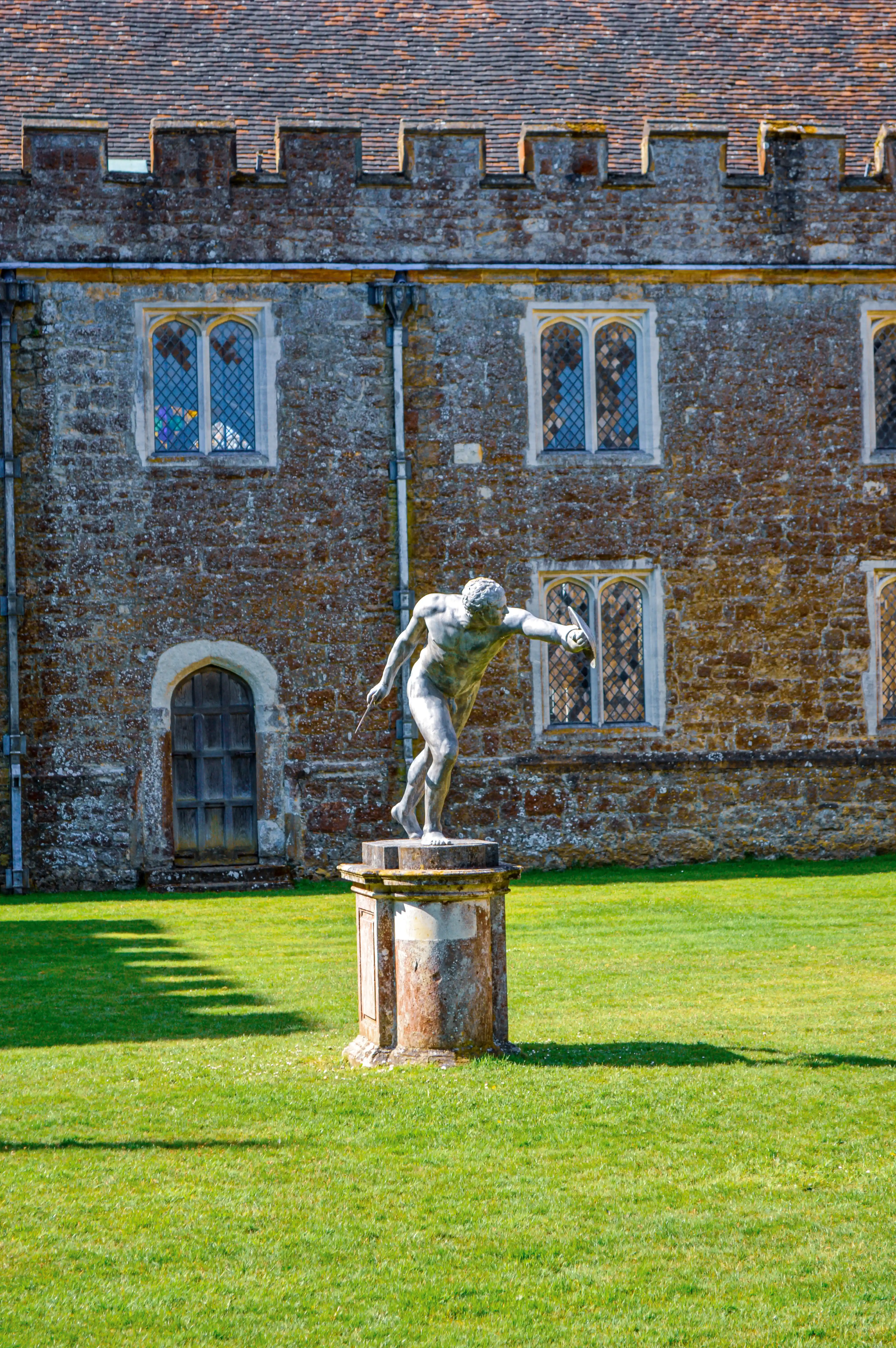 A bronze statue of a muscular male figure, poised as if in mid-action with a spear and shield, stands on a weathered stone plinth in a lush green garden. Behind the statue, a large stone building with battlements, leaded glass windows, and a red tiled roof is visible.
