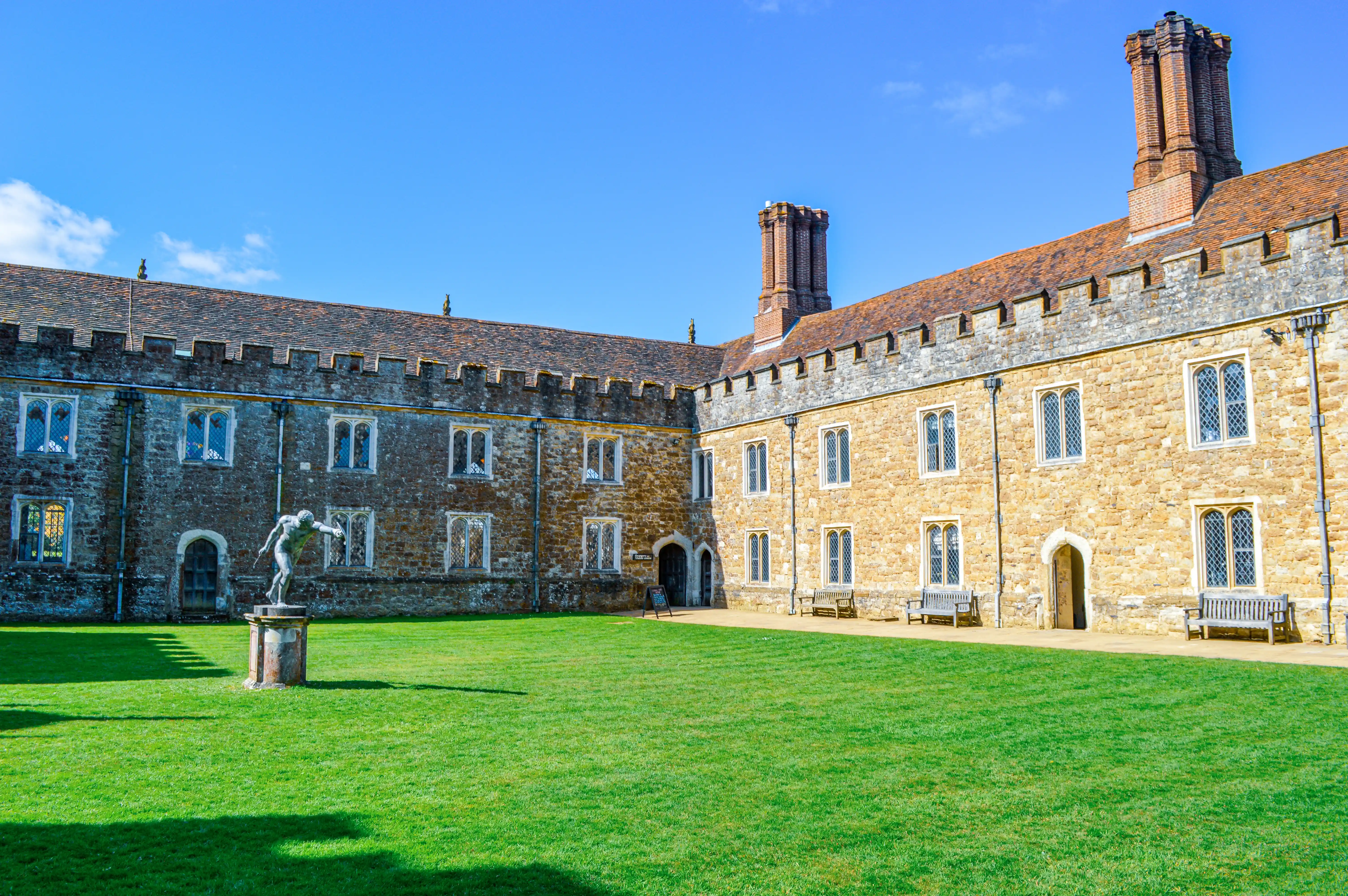 A wide shot of a tranquil courtyard with manicured green grass, an ancient stone building with ornate windows and crenellations, and a bronze statue of a dynamic figure standing on a stone plinth.