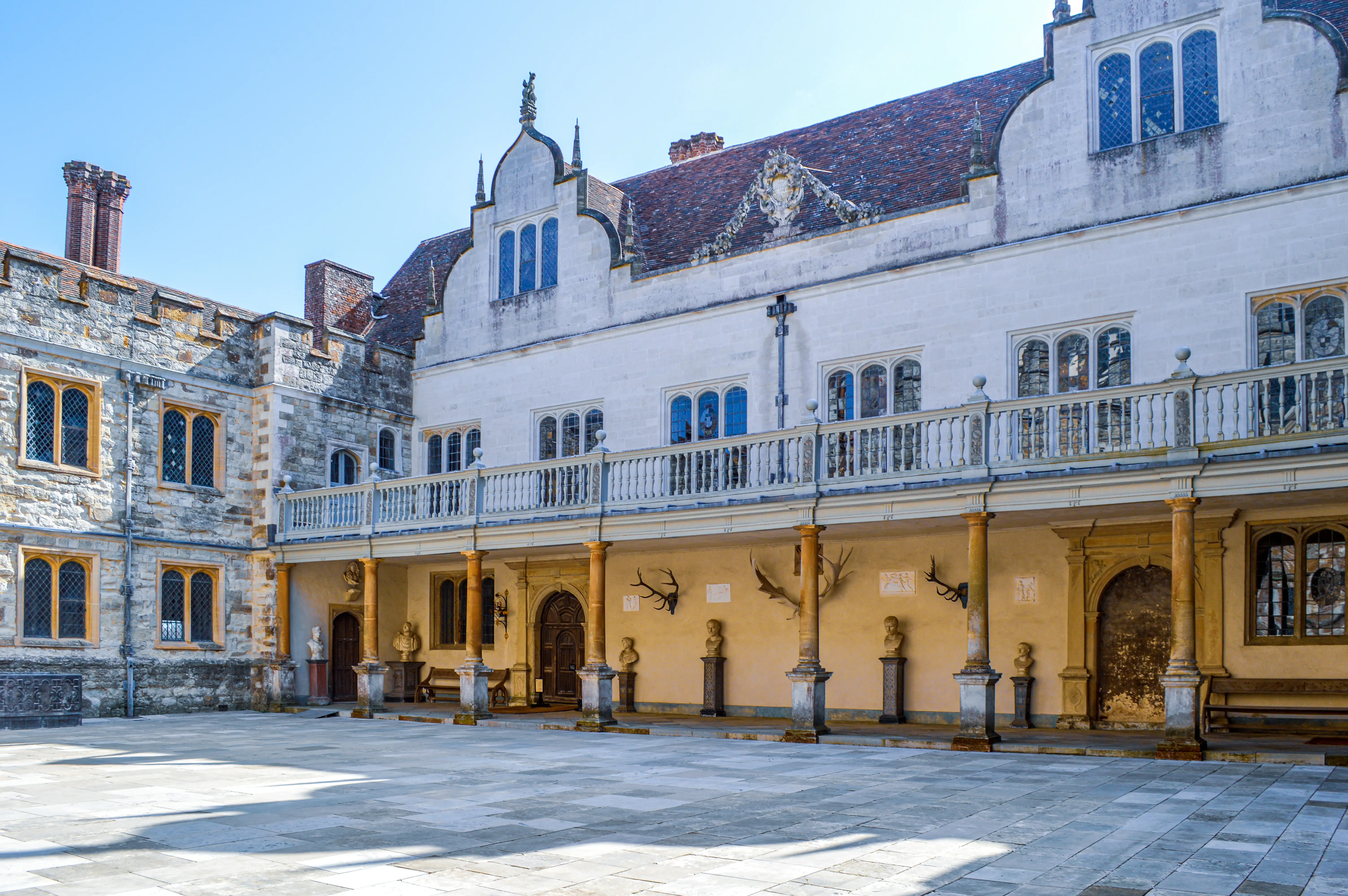 A wide-angle view of a stone-paved courtyard in front of a historic building with a white facade and a stone wing. The white facade has a covered walkway with columns and a decorative balcony above. The stone wing features mullioned windows. The sky is clear and blue.