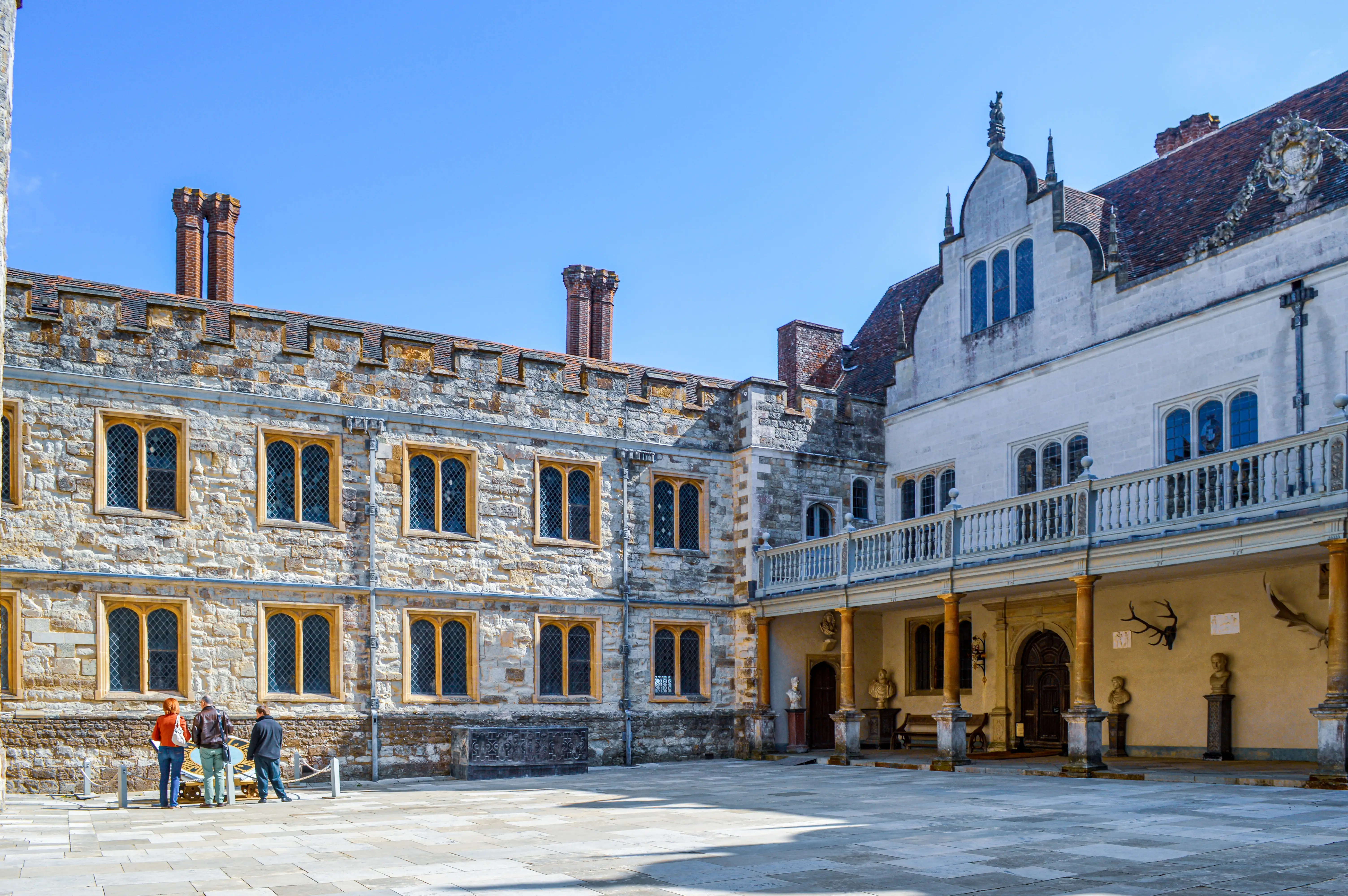 A wide shot of a historic courtyard on a clear day, featuring stone buildings with mullioned windows and a balcony. Three people are standing in the foreground, looking at the buildings.