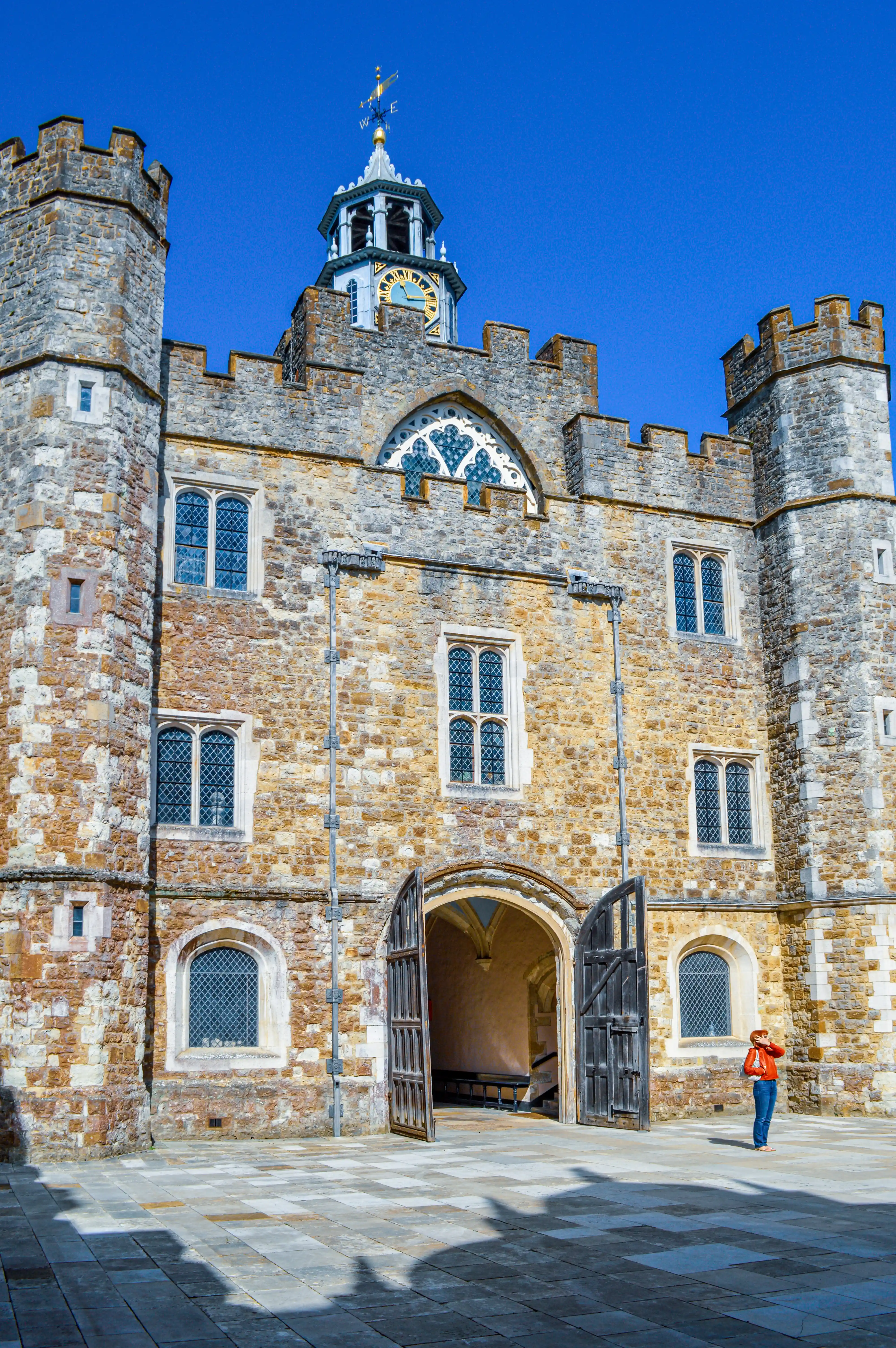 A person in an orange jacket stands in a paved courtyard in front of a large, old stone building with turrets, a clock tower, and open wooden doors.