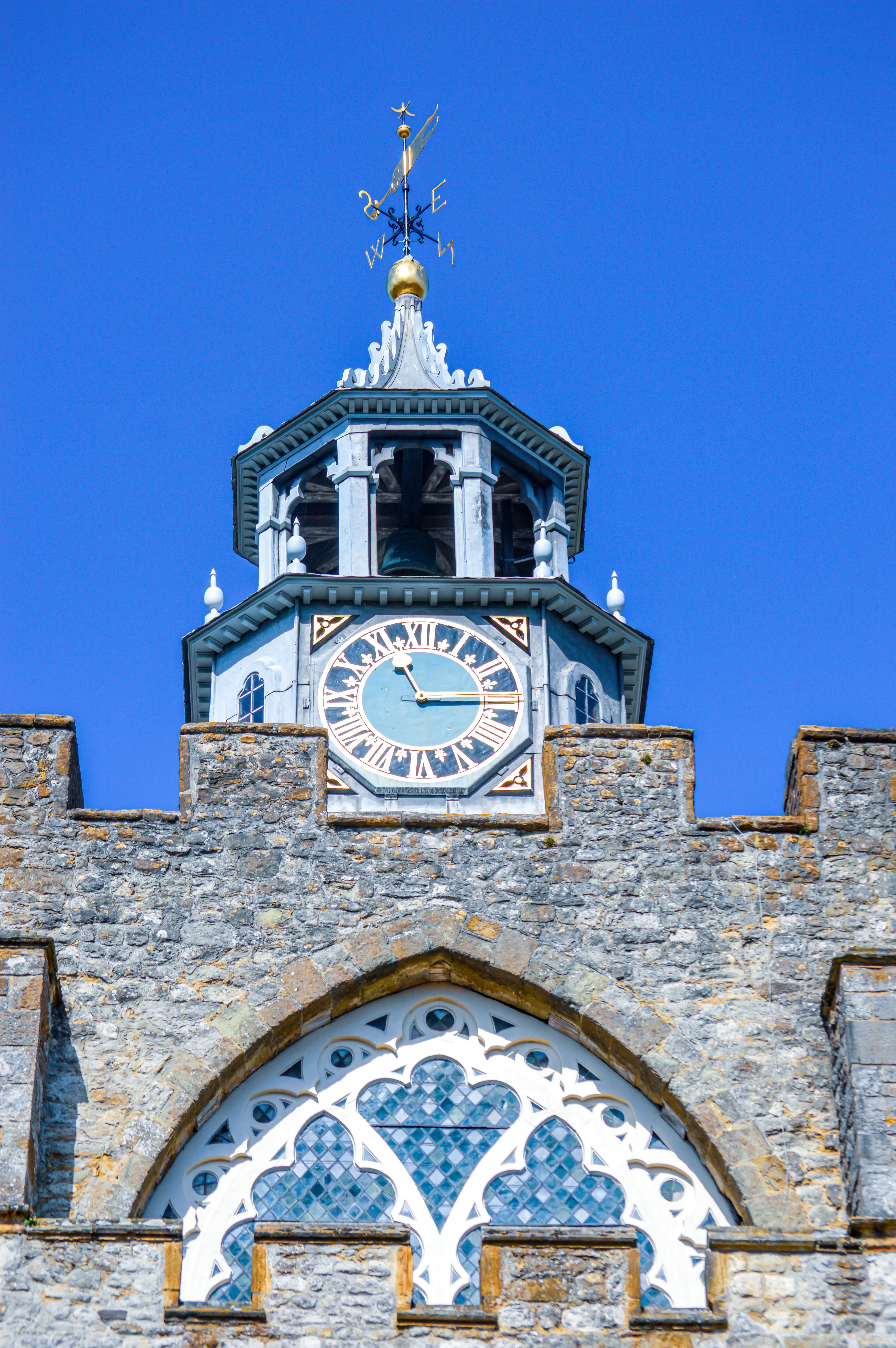 A detailed view of a historic clock tower featuring a large blue clock face with Roman numerals, a bell, a golden weather vane, and rough stone masonry, set against a vivid blue sky.