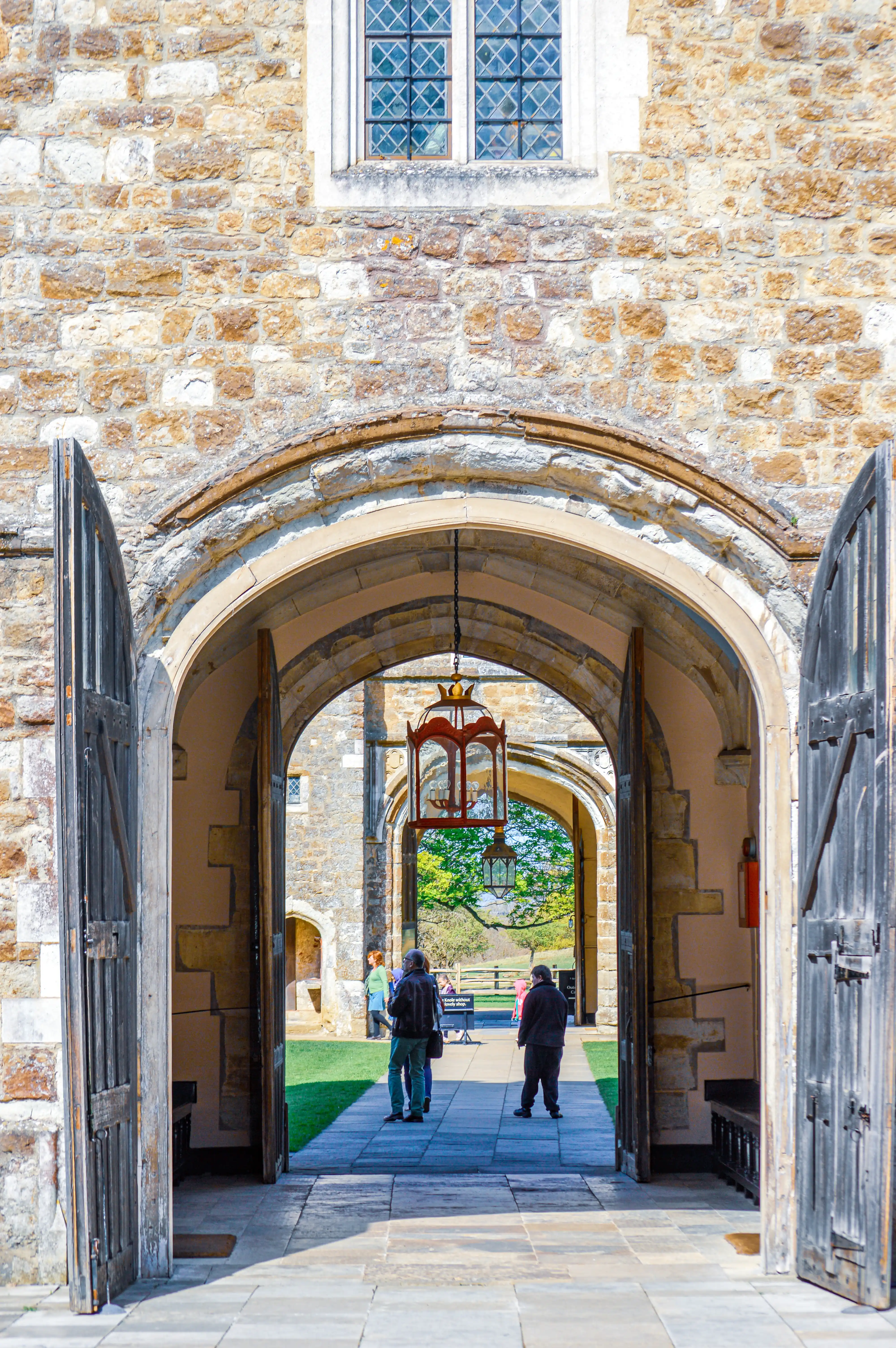 A view through an ancient stone archway into a sunlit courtyard, featuring ornate lanterns and people walking.