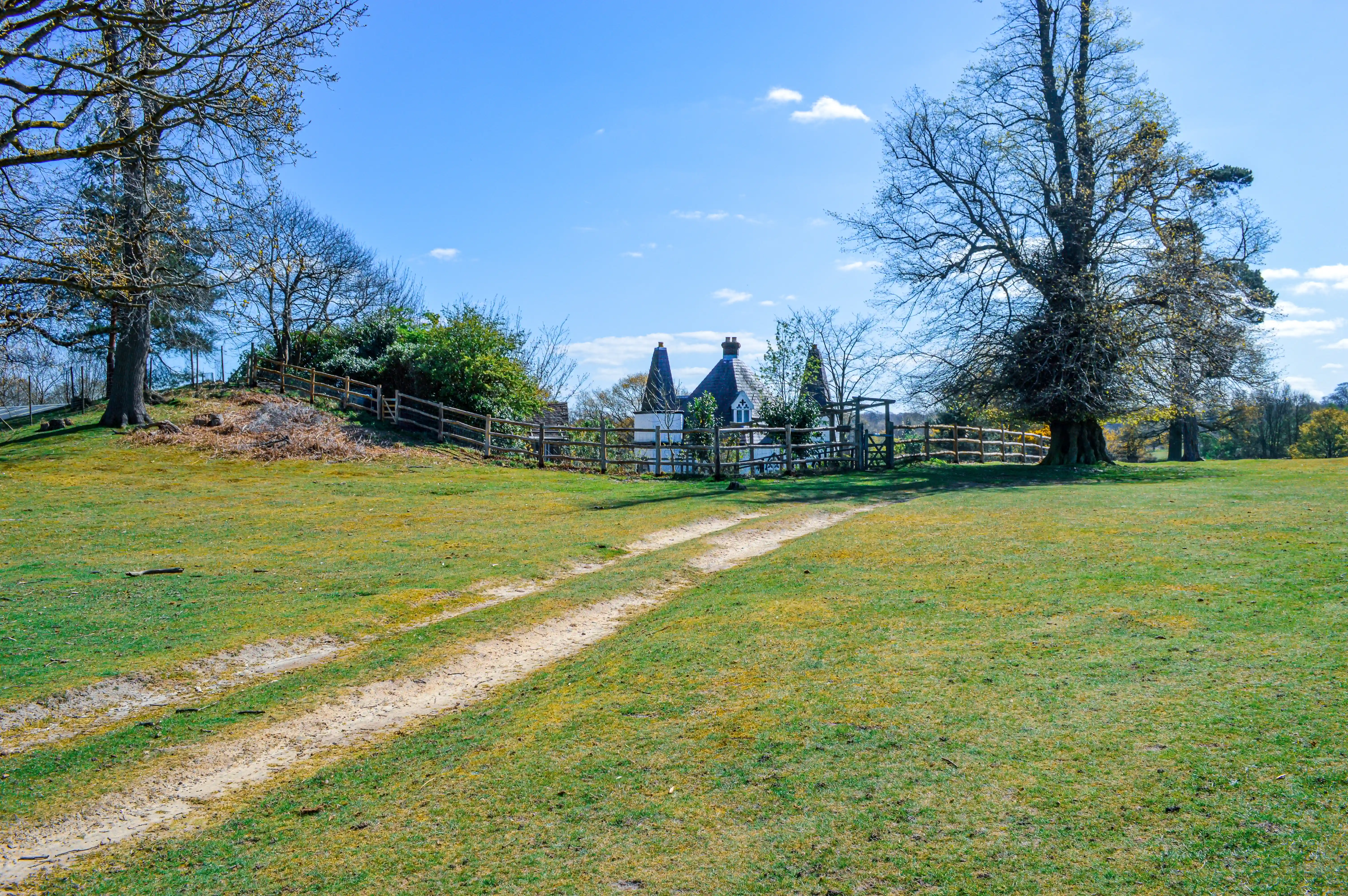 Two dirt tracks lead across a grassy field towards a white oast house with conical roofs, enclosed by a wooden fence, under a bright blue sky with scattered clouds. Large bare trees are visible on either side of the scene.