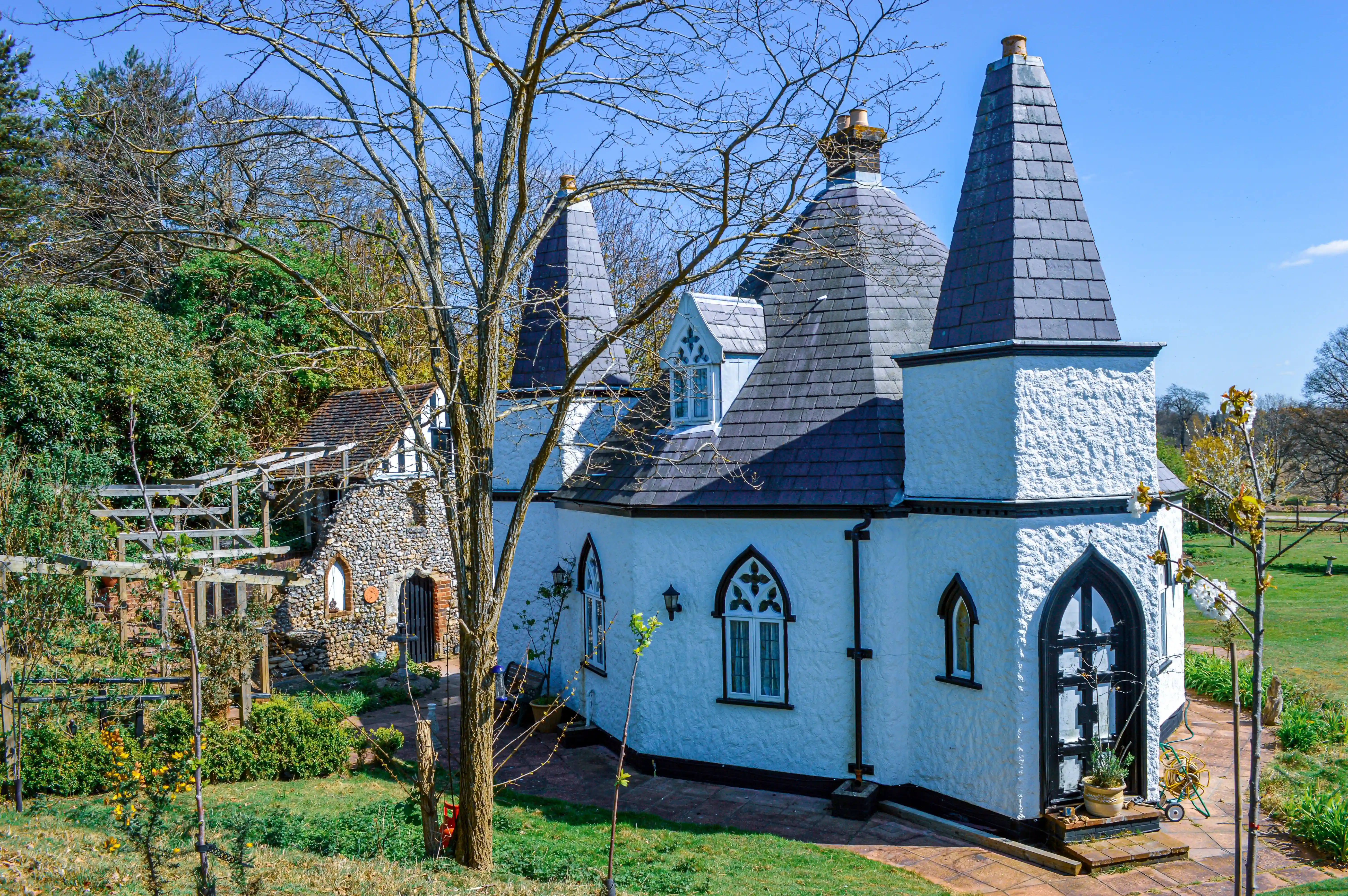 An exterior view of a unique white cottage featuring multiple conical turrets topped with slate tiles, gothic arched windows, and a rough stucco finish. Beside it, a charming, weathered stone structure with a wooden roof stands amidst a garden setting with trees and a bright blue sky.