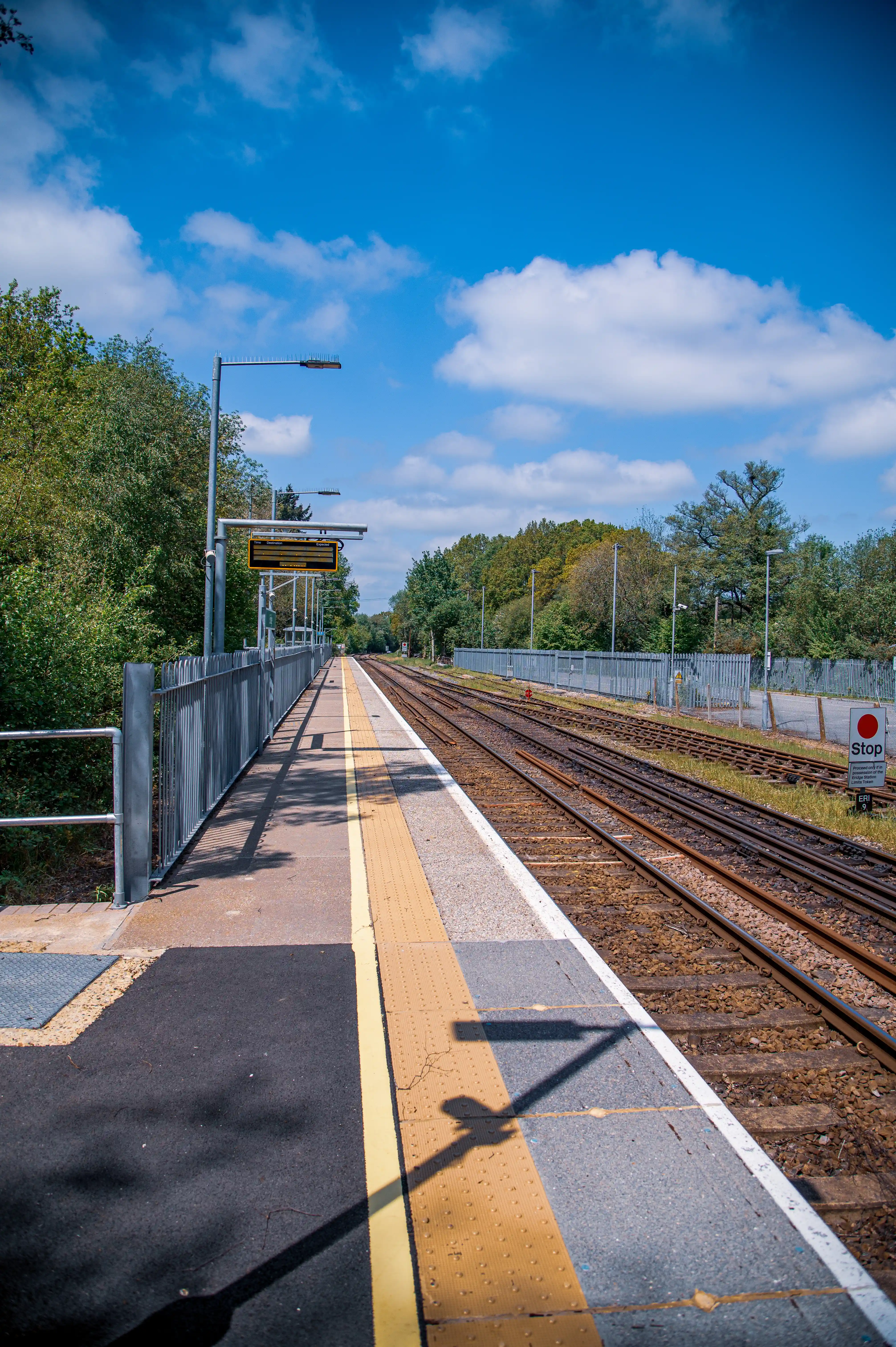 A train station platform with yellow tactile paving and a white line next to multiple railway tracks, under a blue sky with scattered clouds, with trees in the background.