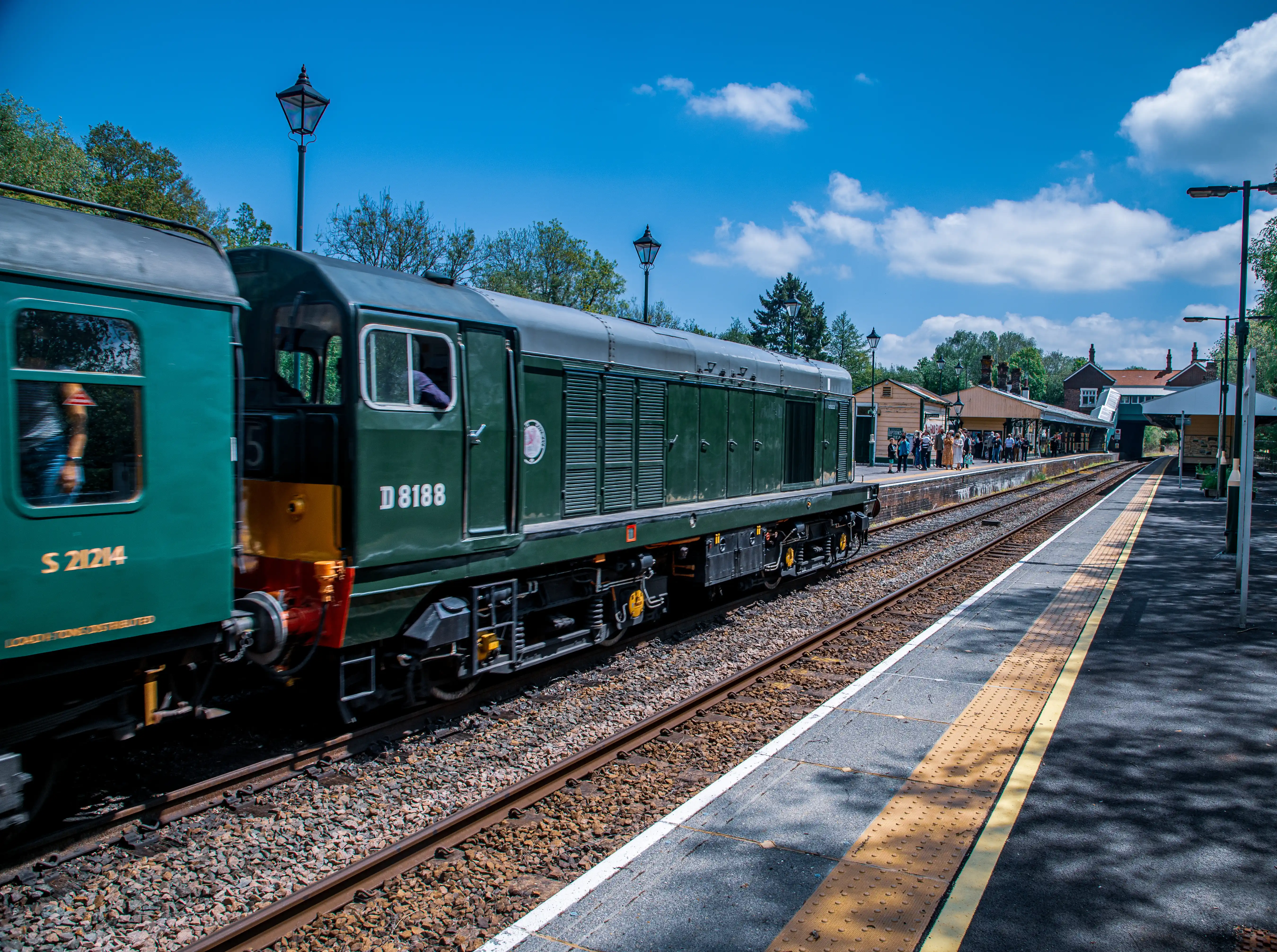 A green diesel locomotive, number D 8188, arriving at a train station platform on a sunny day. Passengers are visible on the platform, and the train has carriage number S 21214.