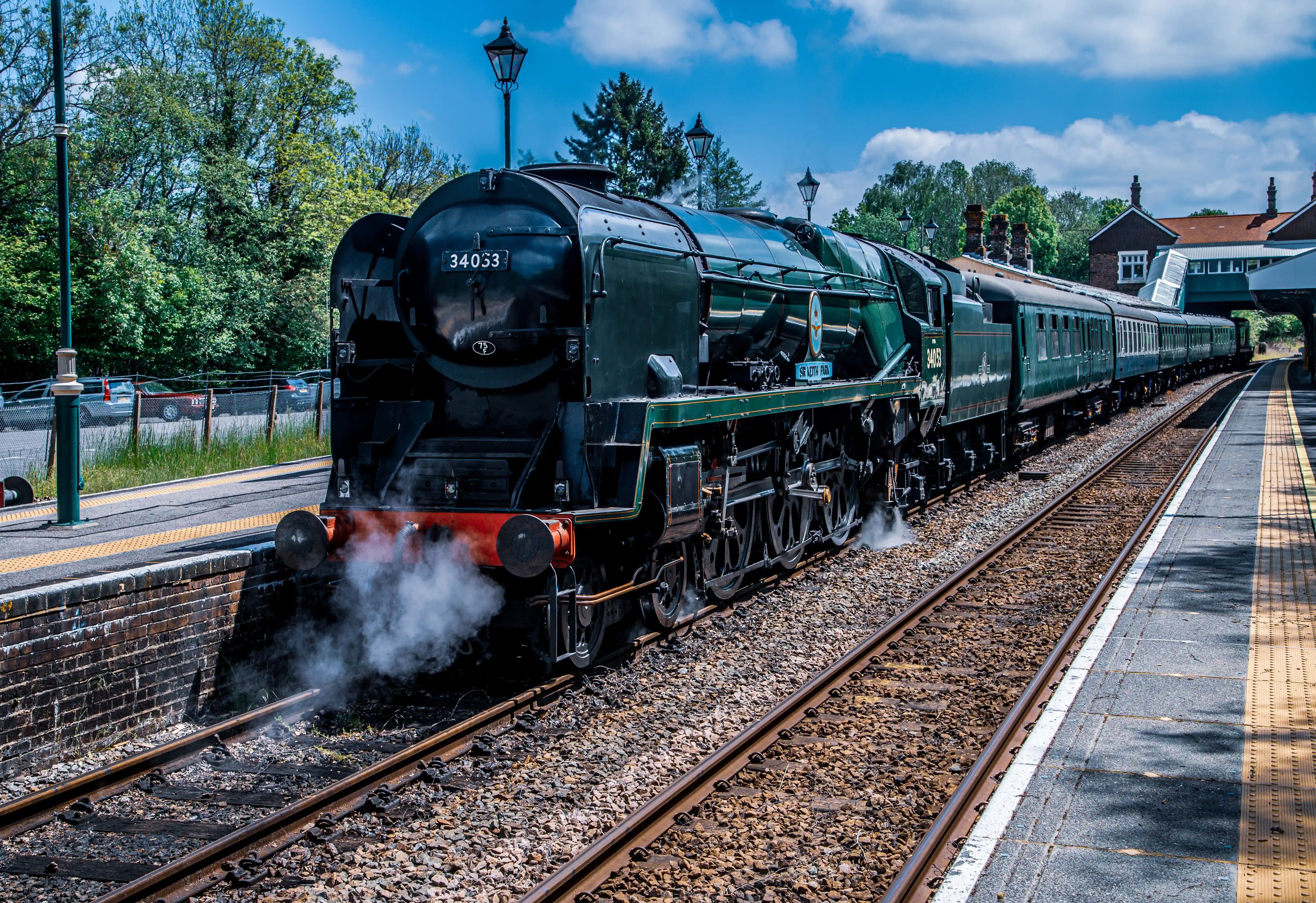 A dark green British steam locomotive with the number 34053 visible on its front is positioned at a train station platform. Steam is emanating from the front of the engine. The train is a long line of carriages extending into the distance, next to a yellow-striped platform. Trees and a building are visible in the background under a cloudy blue sky.