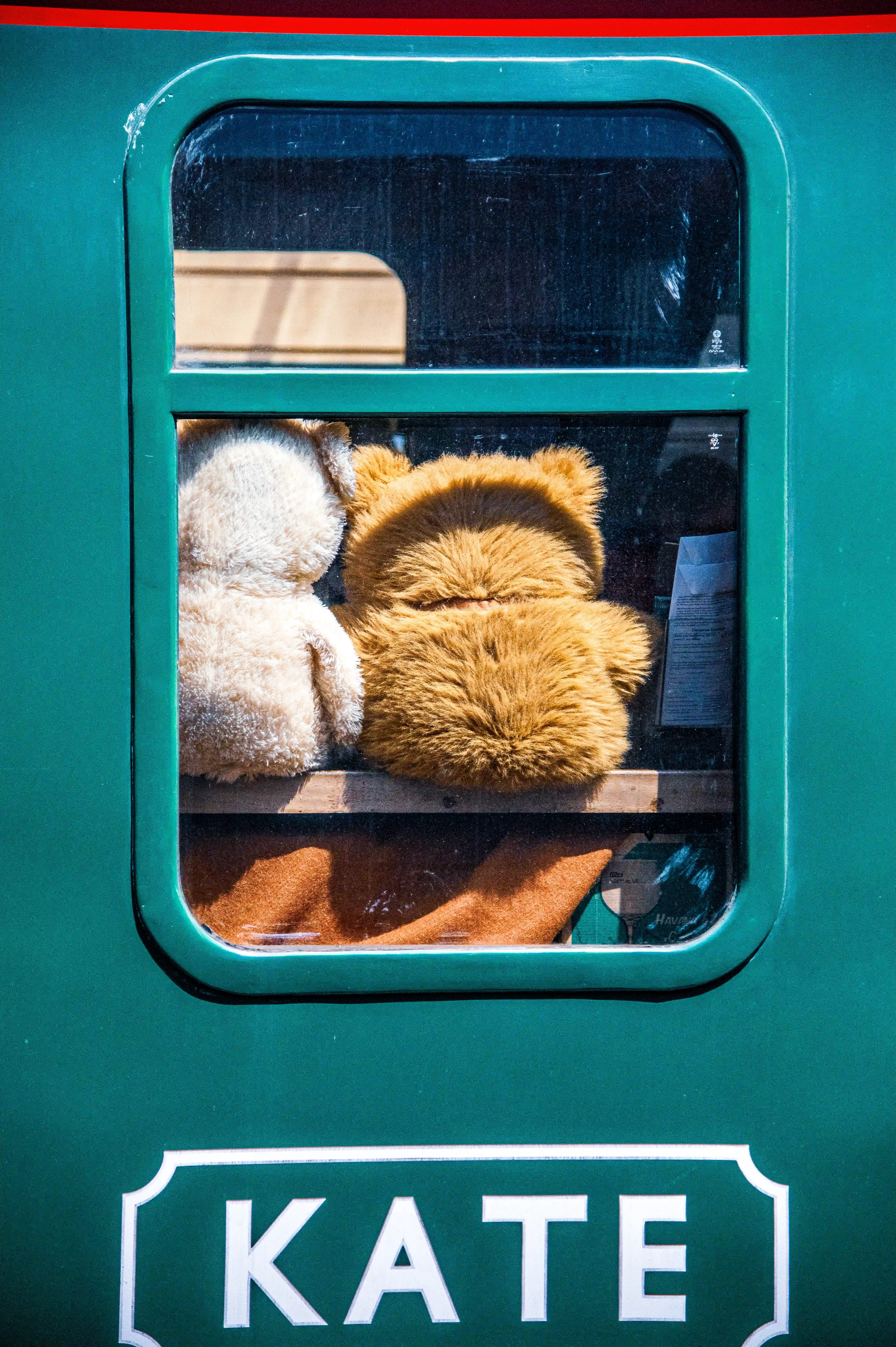 Two teddy bears, one light beige and one golden brown, sit on a shelf inside a green train carriage, viewed through the window. The name KATE is visible on the outside of the train.