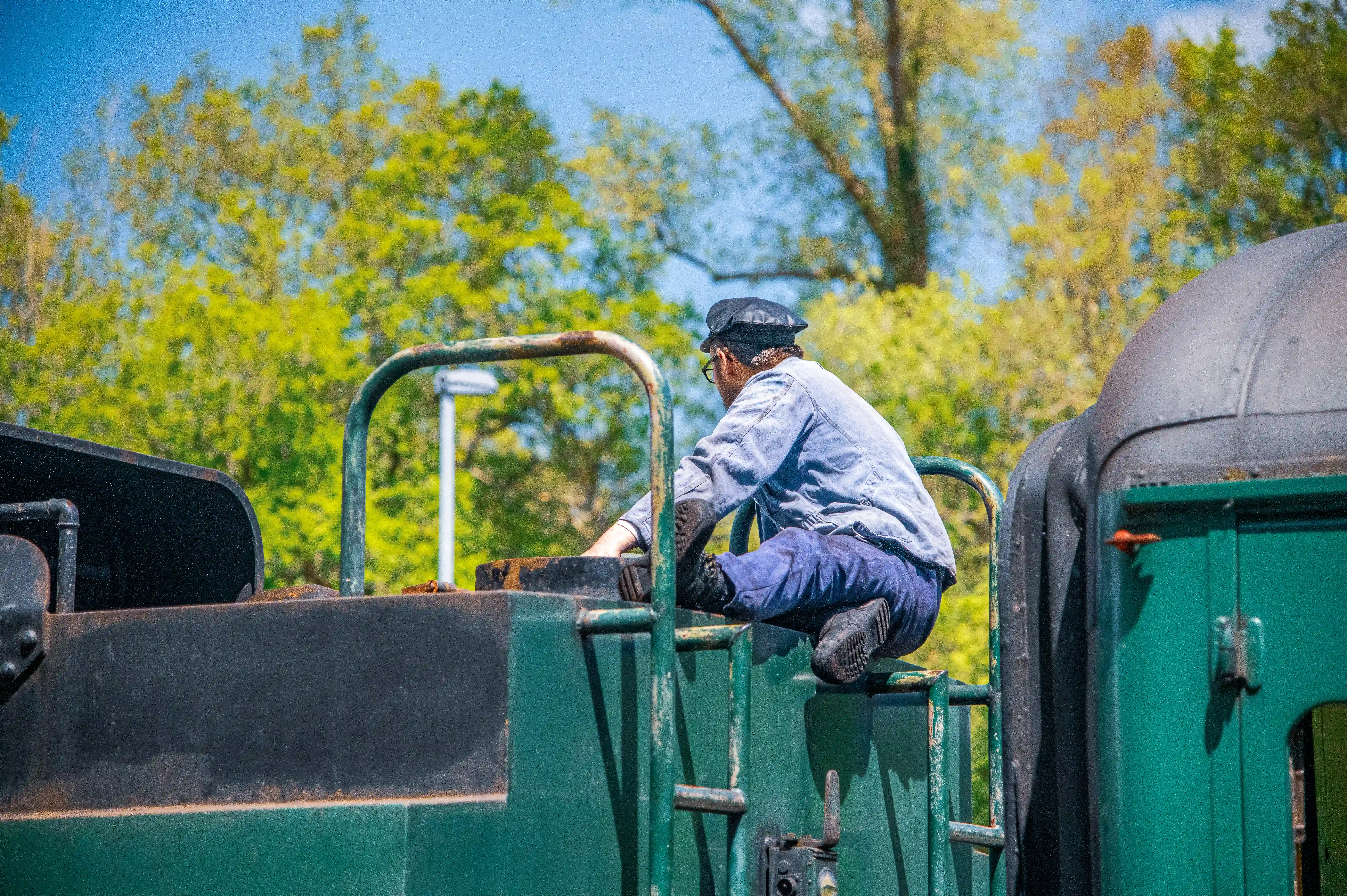 A train conductor wearing a blue shirt, blue trousers, and a flat cap is seated on the exterior of a dark green train. The conductor is leaning forward and appearing to work on the train's machinery. The background consists of green trees under a clear blue sky.