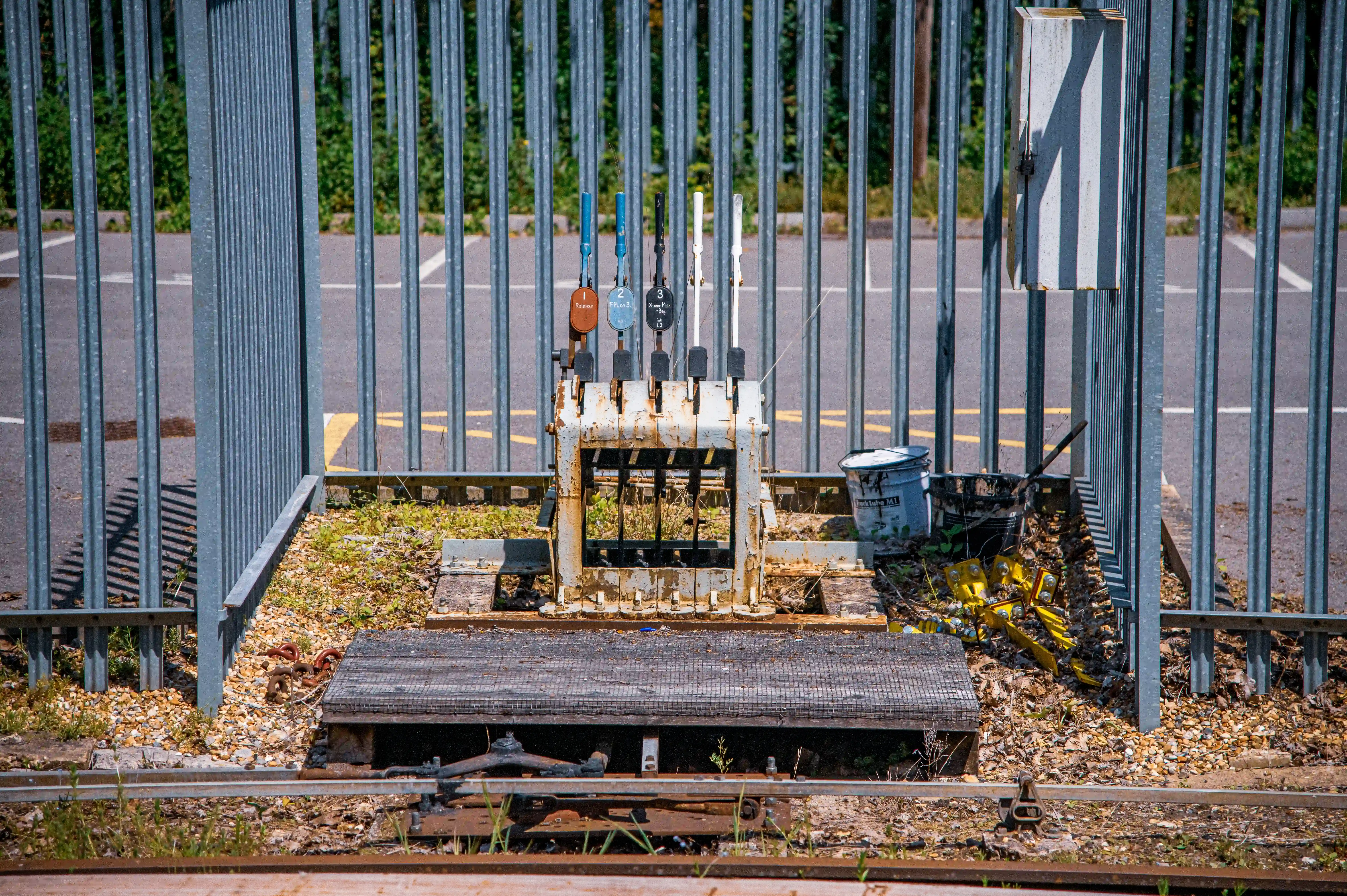 A set of five rusty railway point levers, each with a coloured handle and a numbered plaque, are positioned in front of a grey metal fence. In the foreground are railway tracks and gravel.