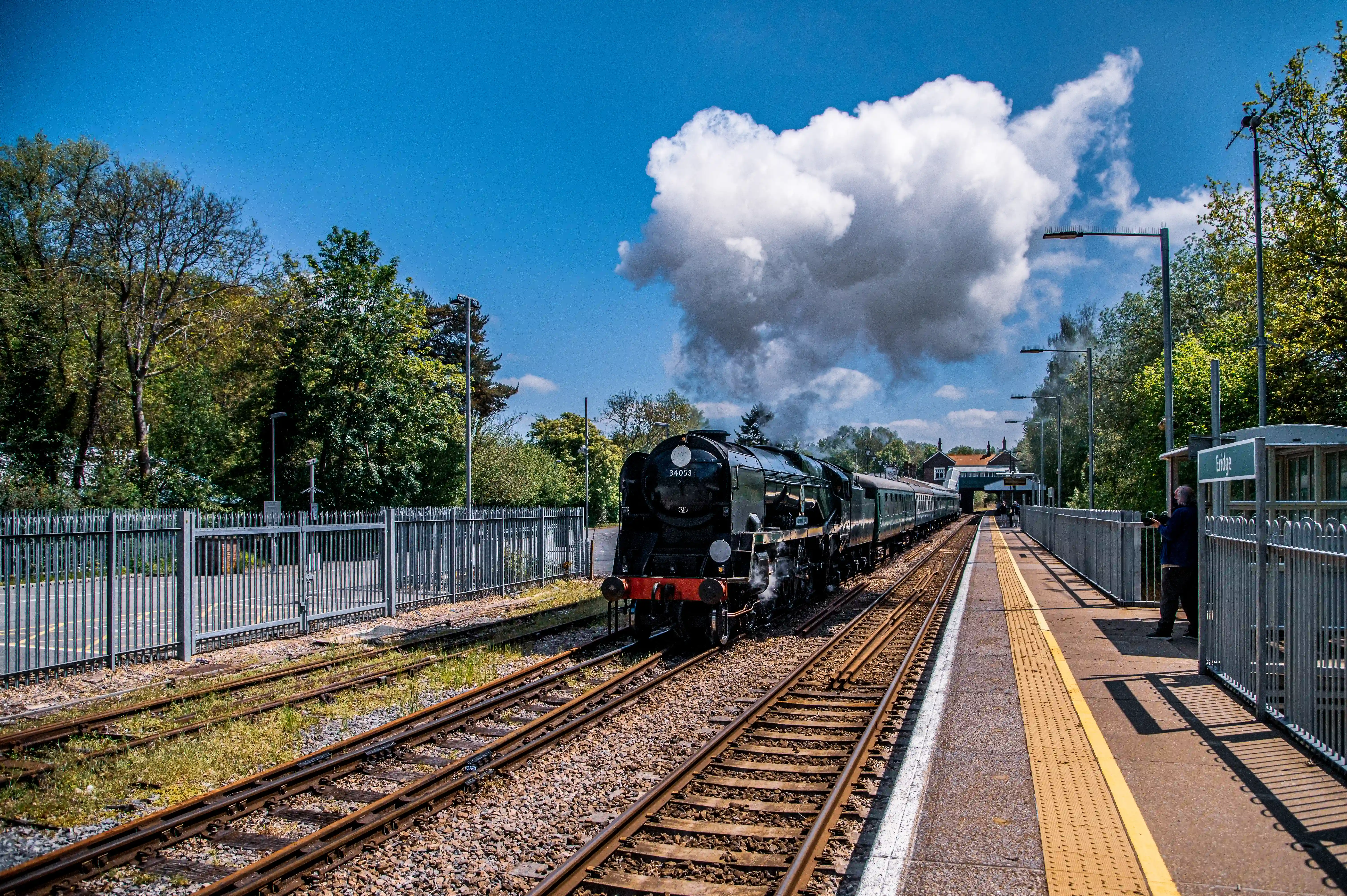A black steam engine, numbered 34053, with carriages attached, is on a railway track at a station platform. There are trees, a shelter, and a bright blue sky with a large cloud.