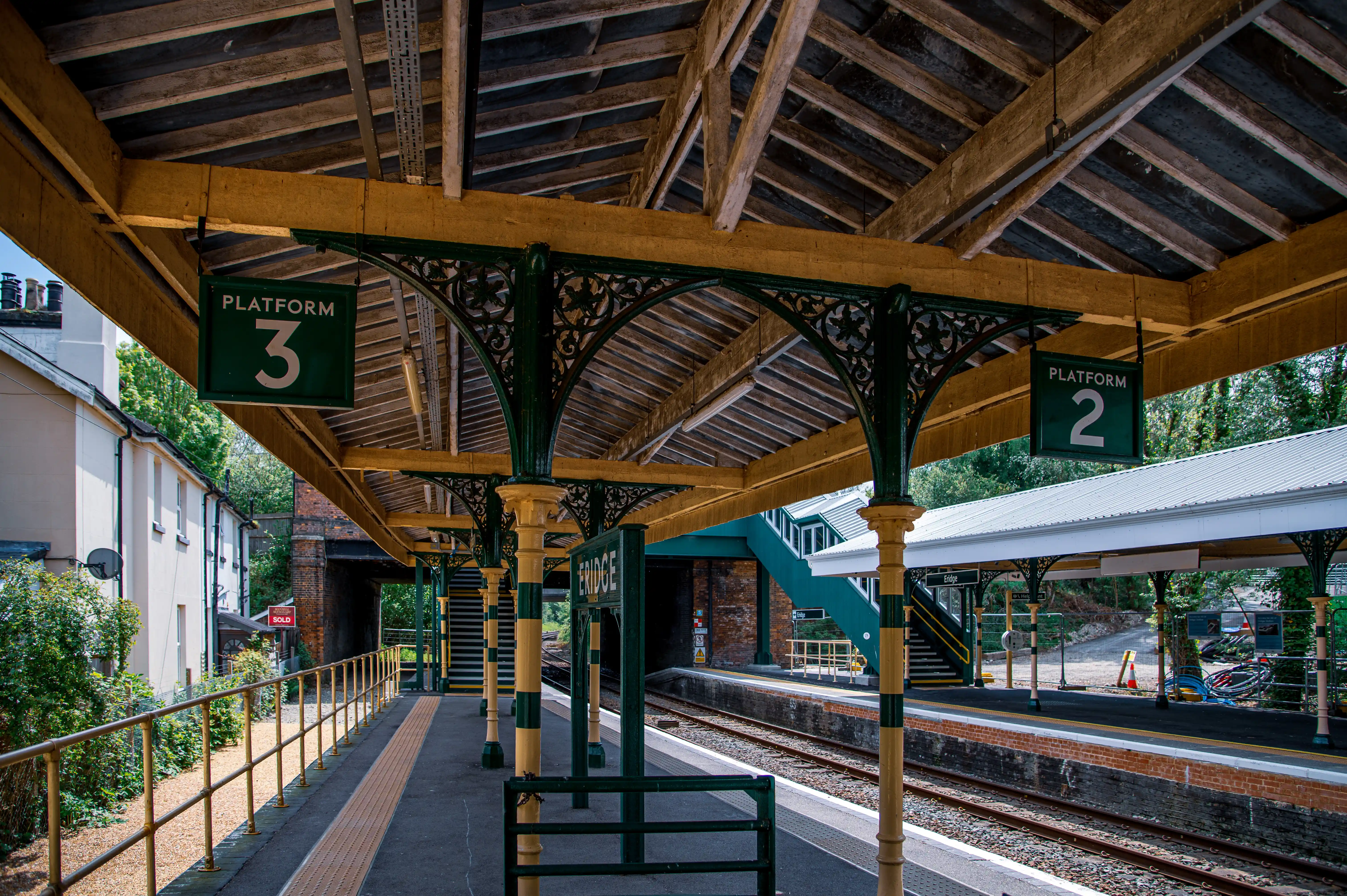 A sunny view of a charming railway station platform with ornate Victorian-era pillars and a wooden roof. Signs indicate PLATFORM 3 and PLATFORM 2, and the station name ERIDGE is visible.