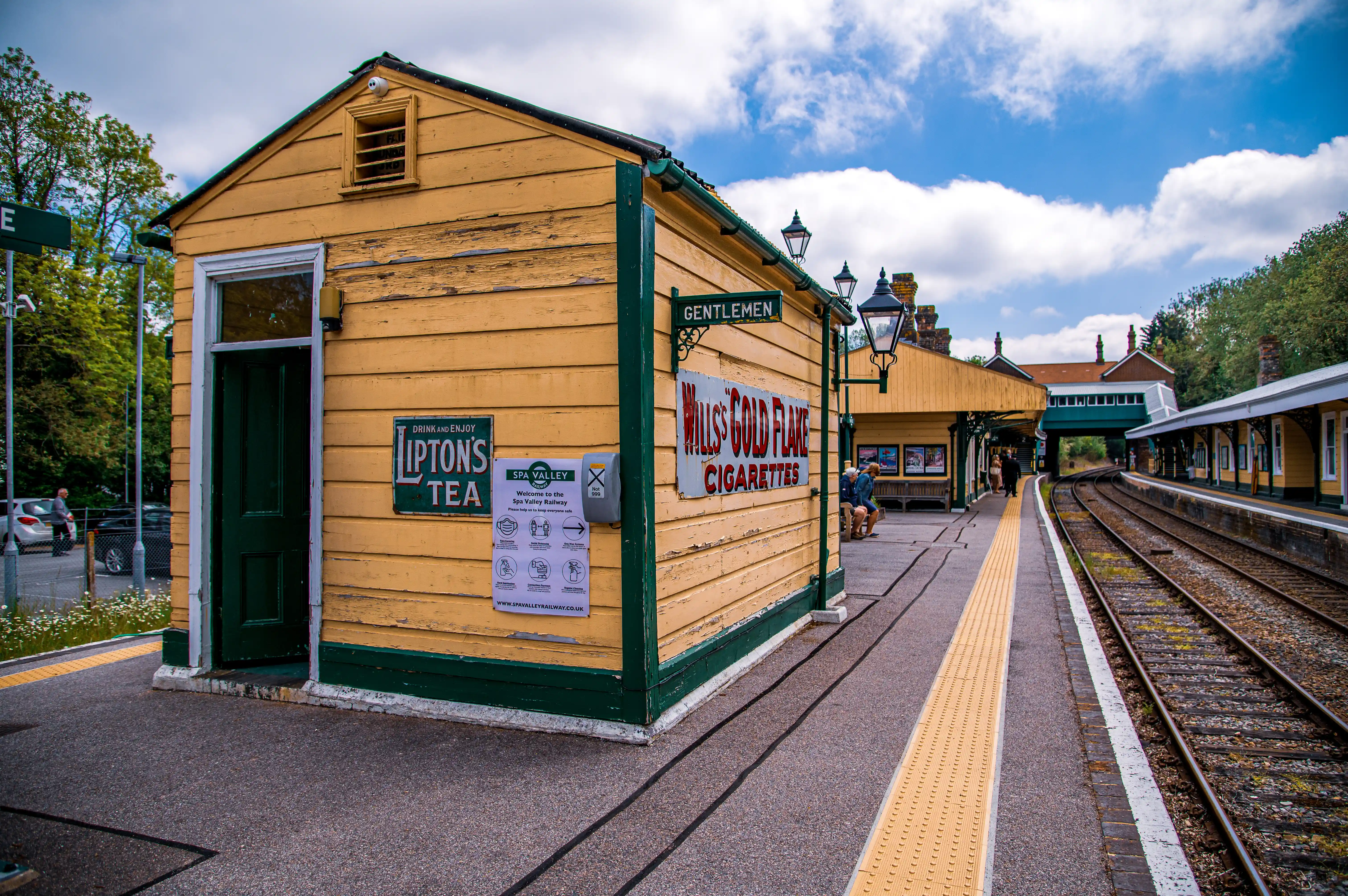 A picturesque view of a traditional British railway station, featuring a quaint, weathered wooden building adorned with vintage advertisements for Lipton's Tea and Wills' Gold Flake Cigarettes. The scene captures the essence of a bygone era, with a platform, railway tracks, and a blue sky dotted with clouds. A sign indicates it's the Spa Valley Railway and another marks a Gentlemen entrance.