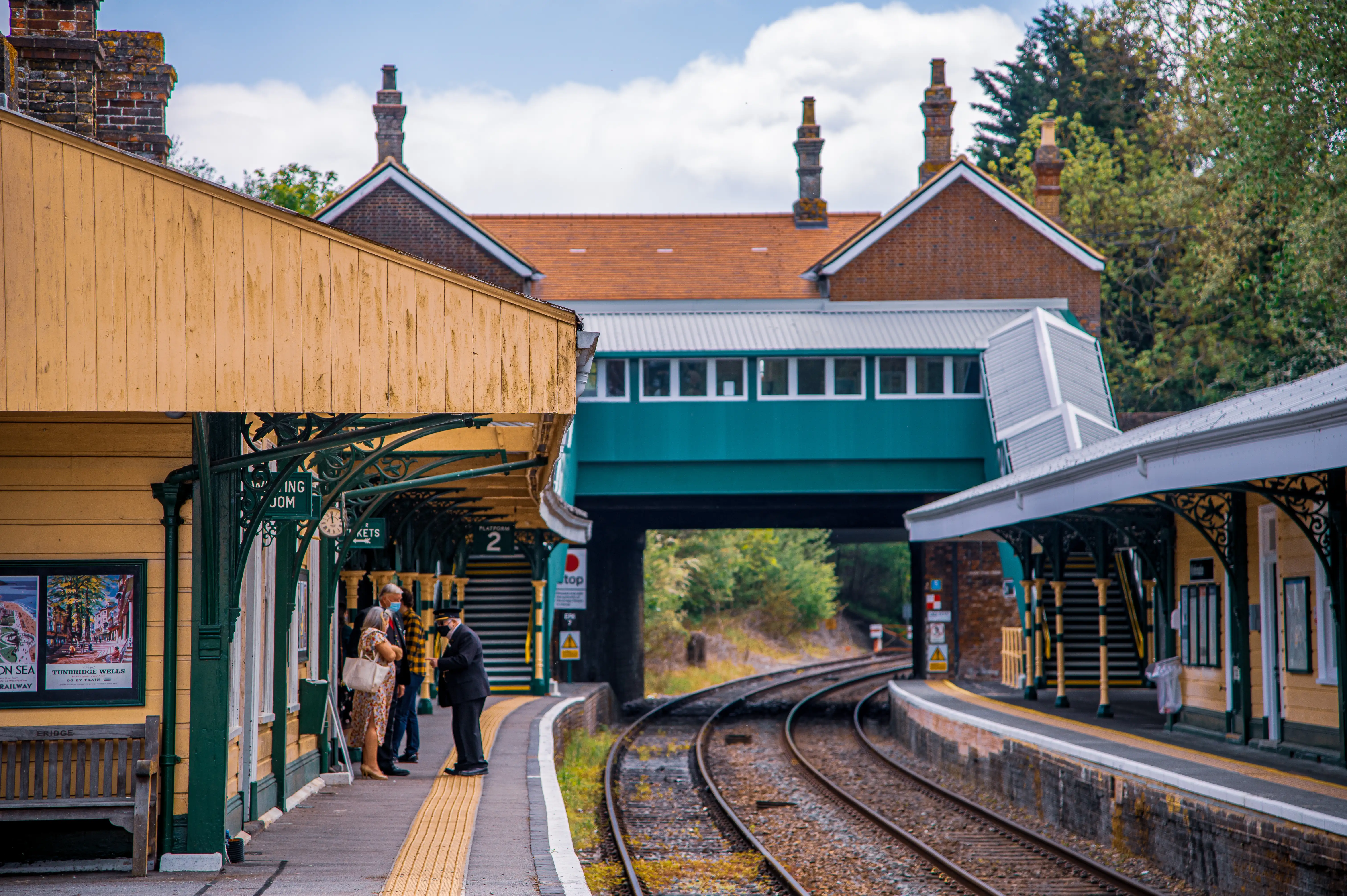 People waiting on a yellow-painted platform at a historic train station with green ornate supports, a teal footbridge crossing train tracks, and brick station buildings.