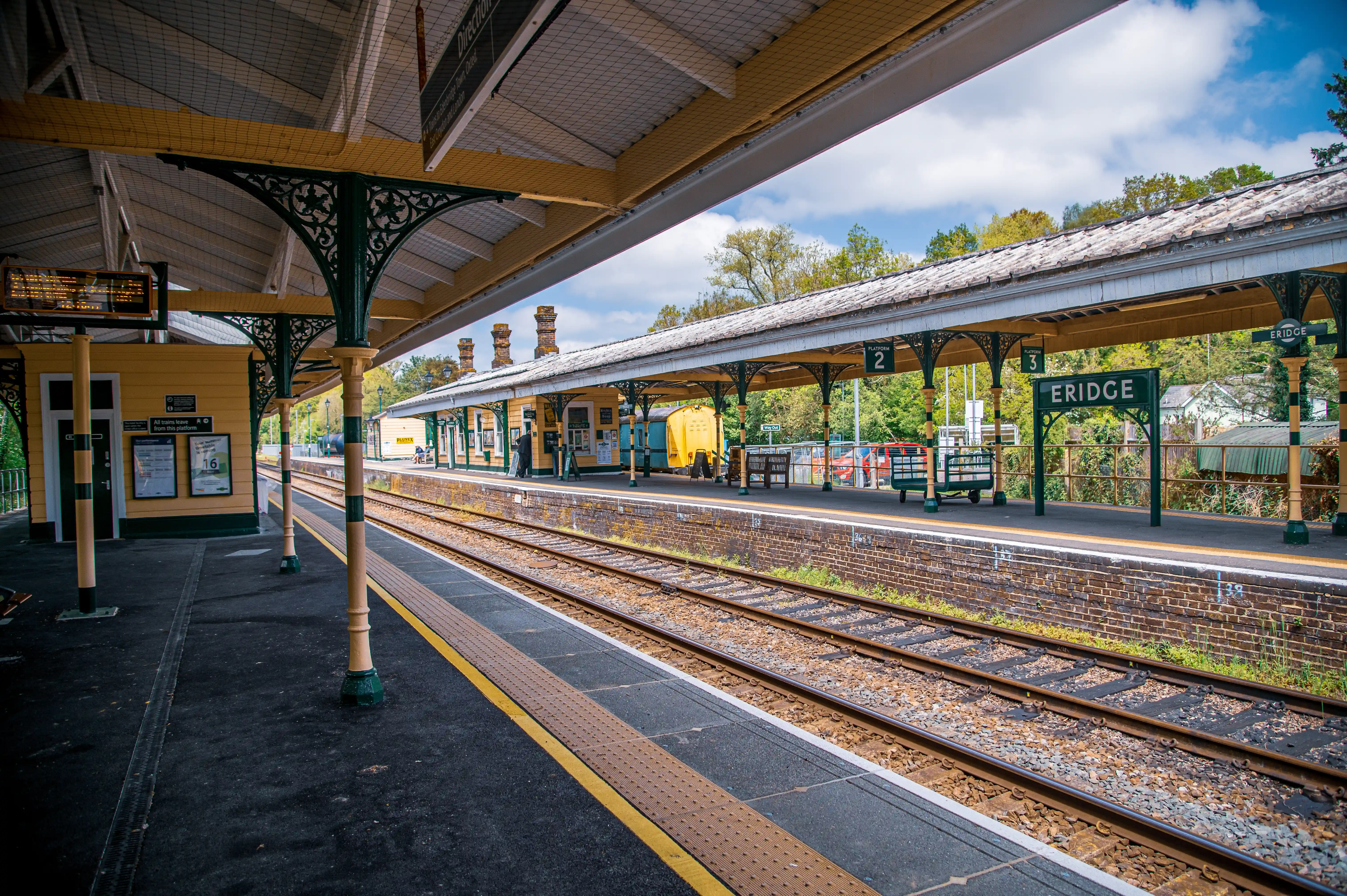 A photograph of Eridge Railway Station platform, featuring train tracks, a yellow and green covered canopy supported by decorative pillars, and station signs including BRIDGE and PLATFORM 2.