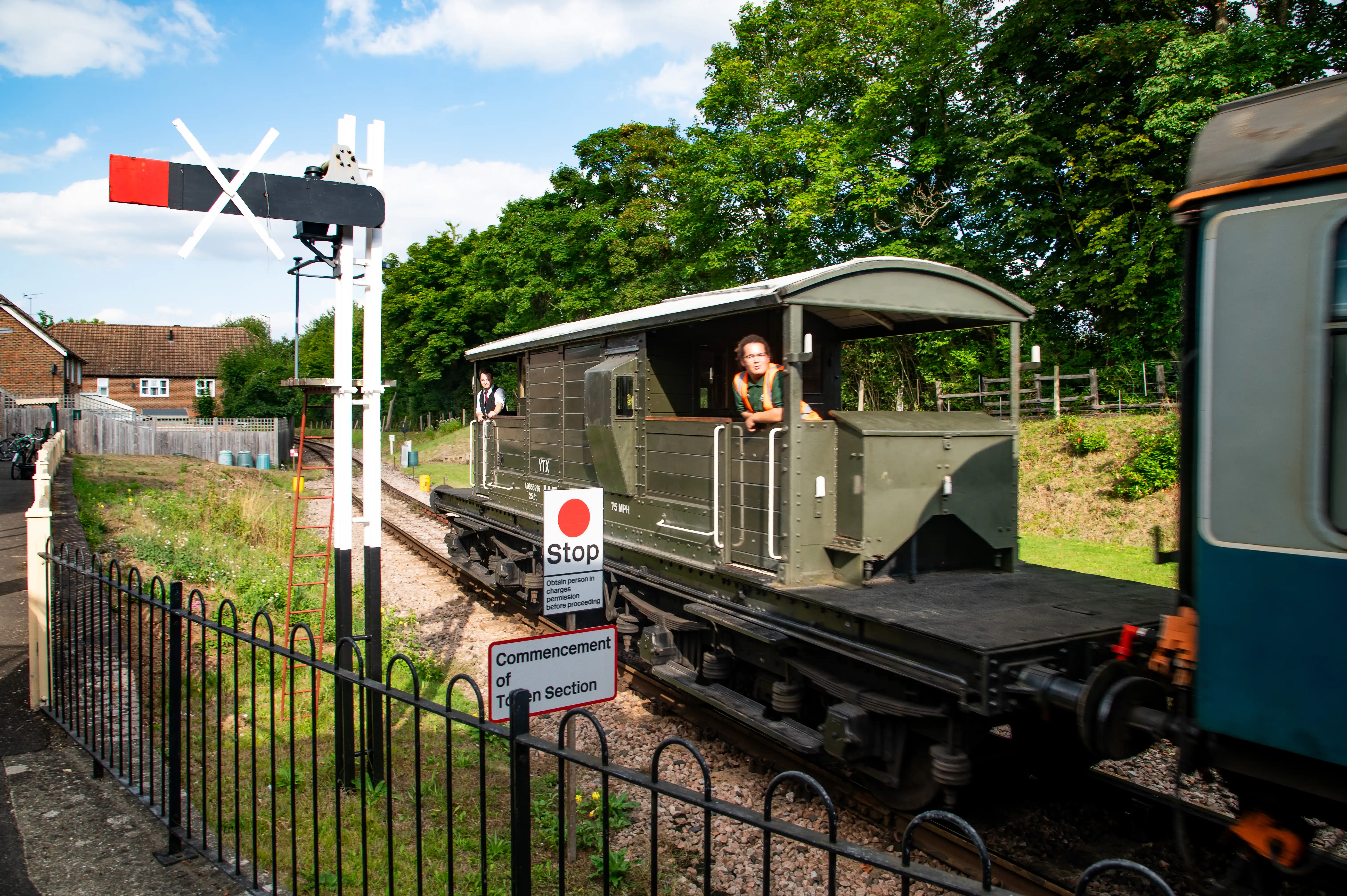 A green vintage train waits at a railway signal on a sunny day. Two people are visible in the open-air carriages. A red and black semaphore signal is raised, and signs reading 