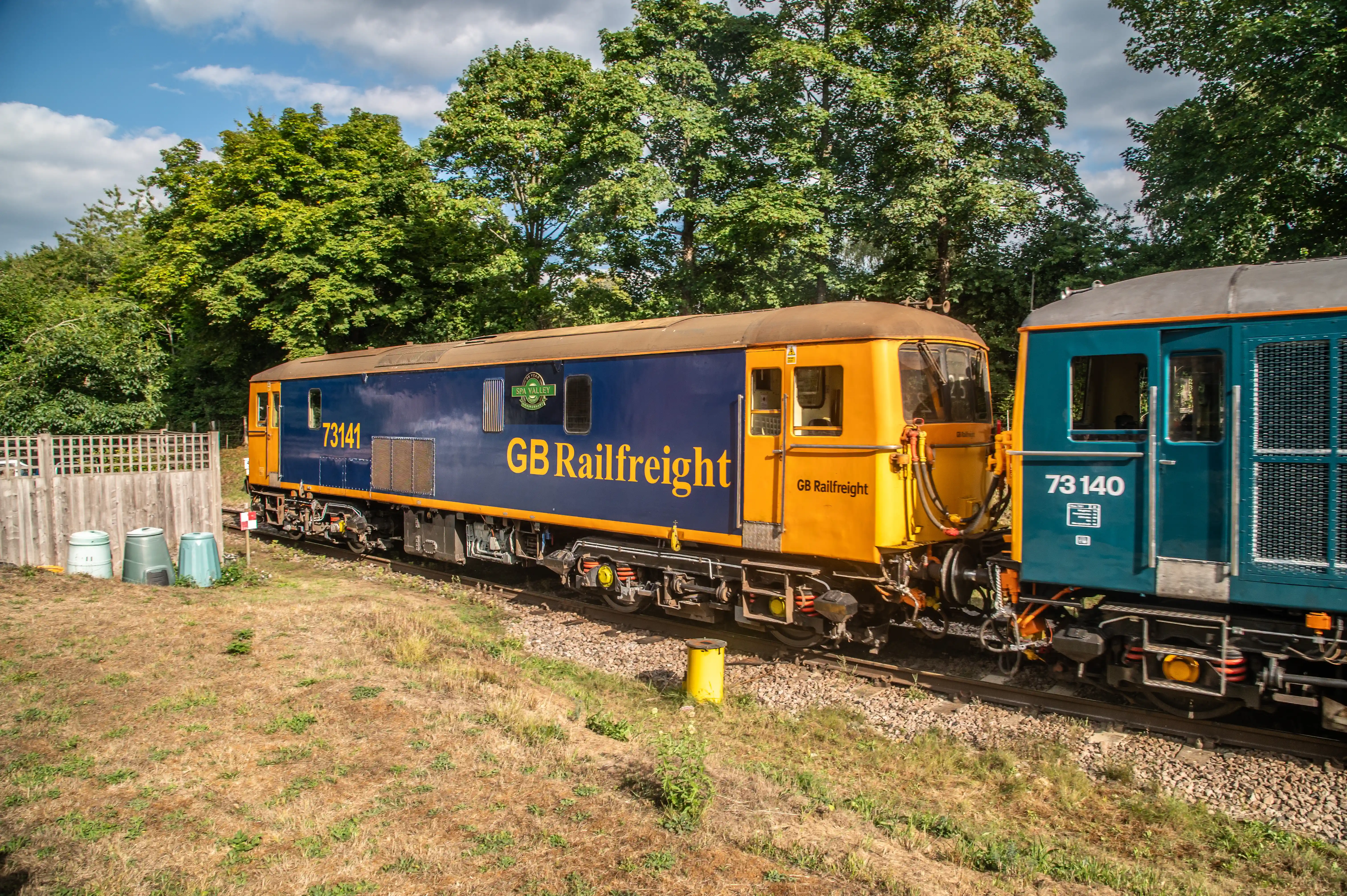 Two GB Railfreight diesel locomotives, one in blue and yellow livery with number 73141 and the other in blue livery with number 73140, stand on a track next to a grassy embankment and trees.