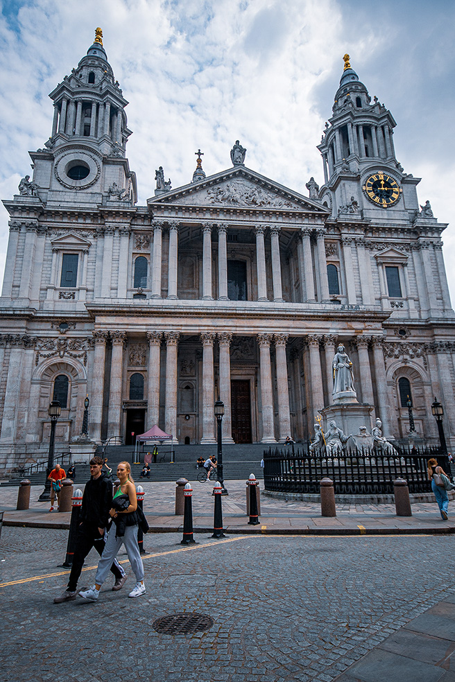 The image depicts St. Paul's Cathedral in London, a renowned architectural landmark. The cathedral features grand columns, intricate stonework, and twin towers with spires. A statue and a clock are prominently displayed on the facade. People are seen walking and standing around the entrance area, indicating it is a popular public space.