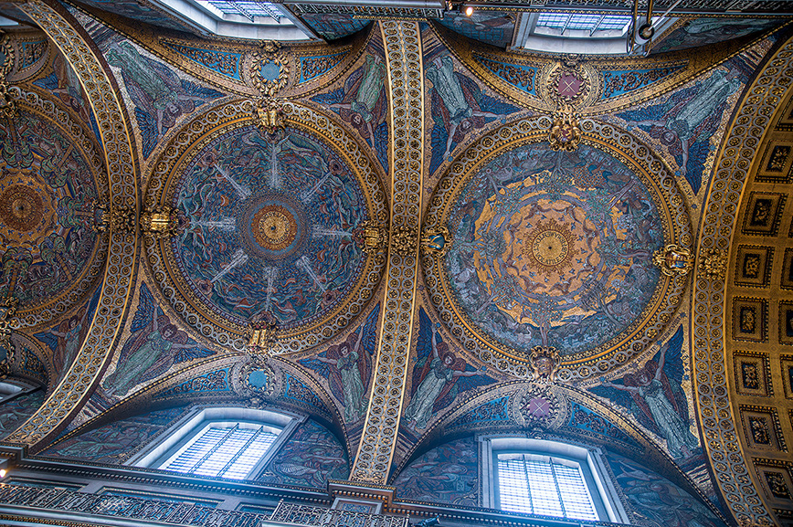 The image depicts an ornately decorated ceiling with intricate patterns and designs. The ceiling features multiple large, circular medallions filled with detailed artwork and surrounded by elaborate borders. The colors predominantly include shades of blue, gold, and red, creating a rich and vibrant visual effect. The architecture includes arched structures and large windows that allow natural light to illuminate the space.