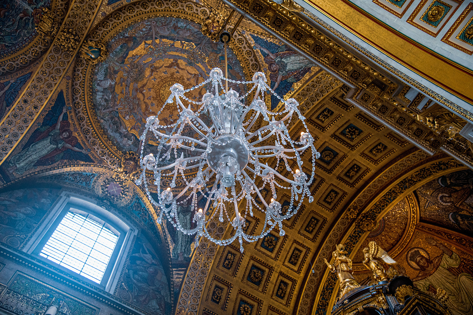 The image depicts the interior of an ornate, historical building, likely a cathedral or palace. The ceiling is richly decorated with intricate frescoes and gold accents. A large, elegant crystal chandelier hangs prominently in the center. The architecture features arched windows and detailed sculptures, contributing to the grandeur of the space.