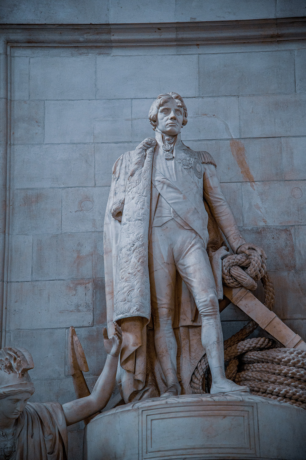 The image depicts a statue of a historical figure dressed in a military uniform, standing confidently with a rope and an oar at his side. The statue is made of stone and is set against a stone wall. The figure is wearing a detailed coat with epaulettes and a sash, suggesting a high rank. The statue appears to be part of a larger monument, with other elements visible at the base.