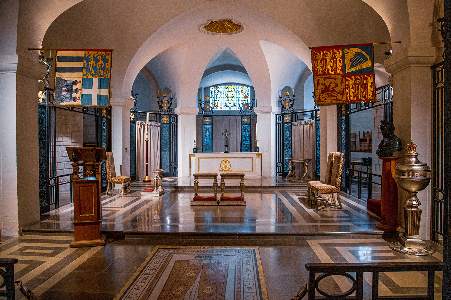The image depicts an interior view of a chapel, showcasing its altar, stained glass windows, and various religious artifacts. The chapel features ornate decorations, including banners with heraldic symbols, a polished floor with intricate designs, and wooden furniture such as chairs and a pulpit. The altar is centrally placed, adorned with a cross and surrounded by candles. The overall ambiance is serene and reverent, indicative of a place of worship.