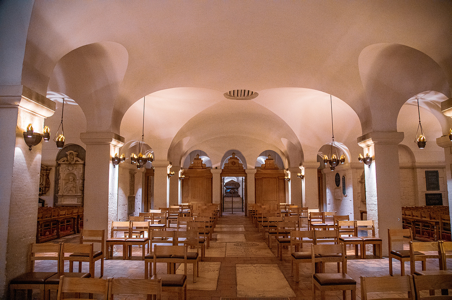 The image depicts an interior view of a historical or religious building, likely a church or cathedral. The space features arched ceilings supported by columns, with ornate chandeliers hanging from the ceiling. Wooden chairs are arranged in rows, suggesting a seating area for congregants. The walls are adorned with plaques and possibly religious artwork. The overall ambiance is serene and solemn, indicative of a place of worship.
