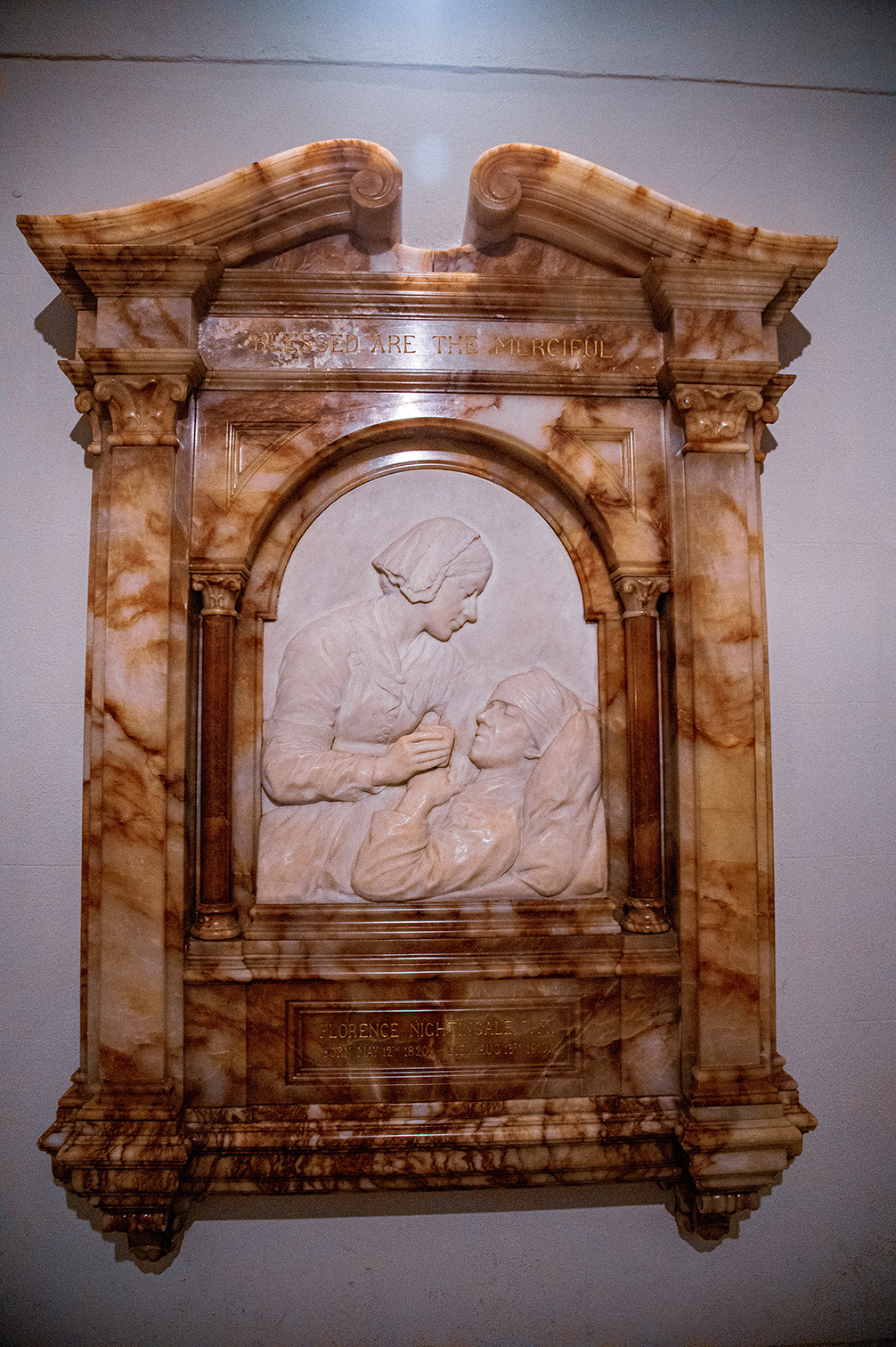 The image depicts a memorial sculpture dedicated to Florence Nightingale, featuring her in a caring pose with a wounded soldier. The sculpture is framed by an ornate, classical-style marble structure with the inscription 'Blessed are the Merciful' at the top. The base of the sculpture includes a plaque with her name and dates of birth and death.