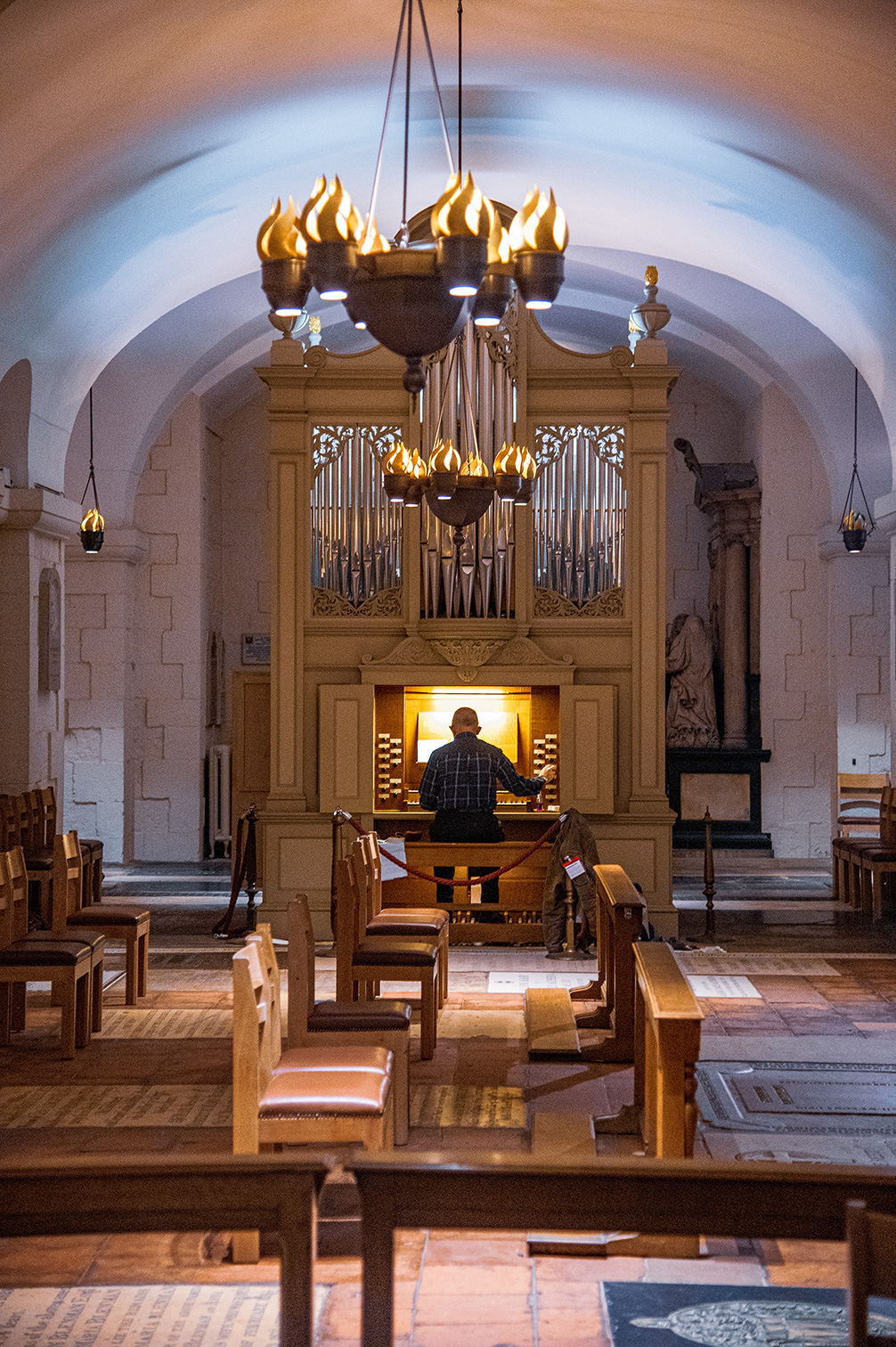 The image depicts the interior of a church, focusing on an organist playing an organ. The church features wooden pews, a large pipe organ with a wooden casing, and several chandeliers with lit candles. The architecture includes arched ceilings and stone walls, creating a serene and reverent atmosphere.