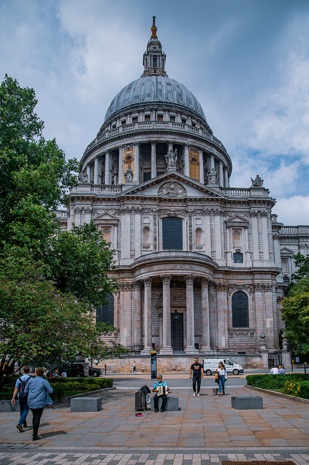 The image depicts St. Paul's Cathedral in London, a renowned architectural landmark with its iconic dome and classical facade. In the foreground, people are seen walking and a man is playing an accordion, adding a lively atmosphere to the scene. The cathedral is surrounded by greenery and a few vehicles are parked nearby.
