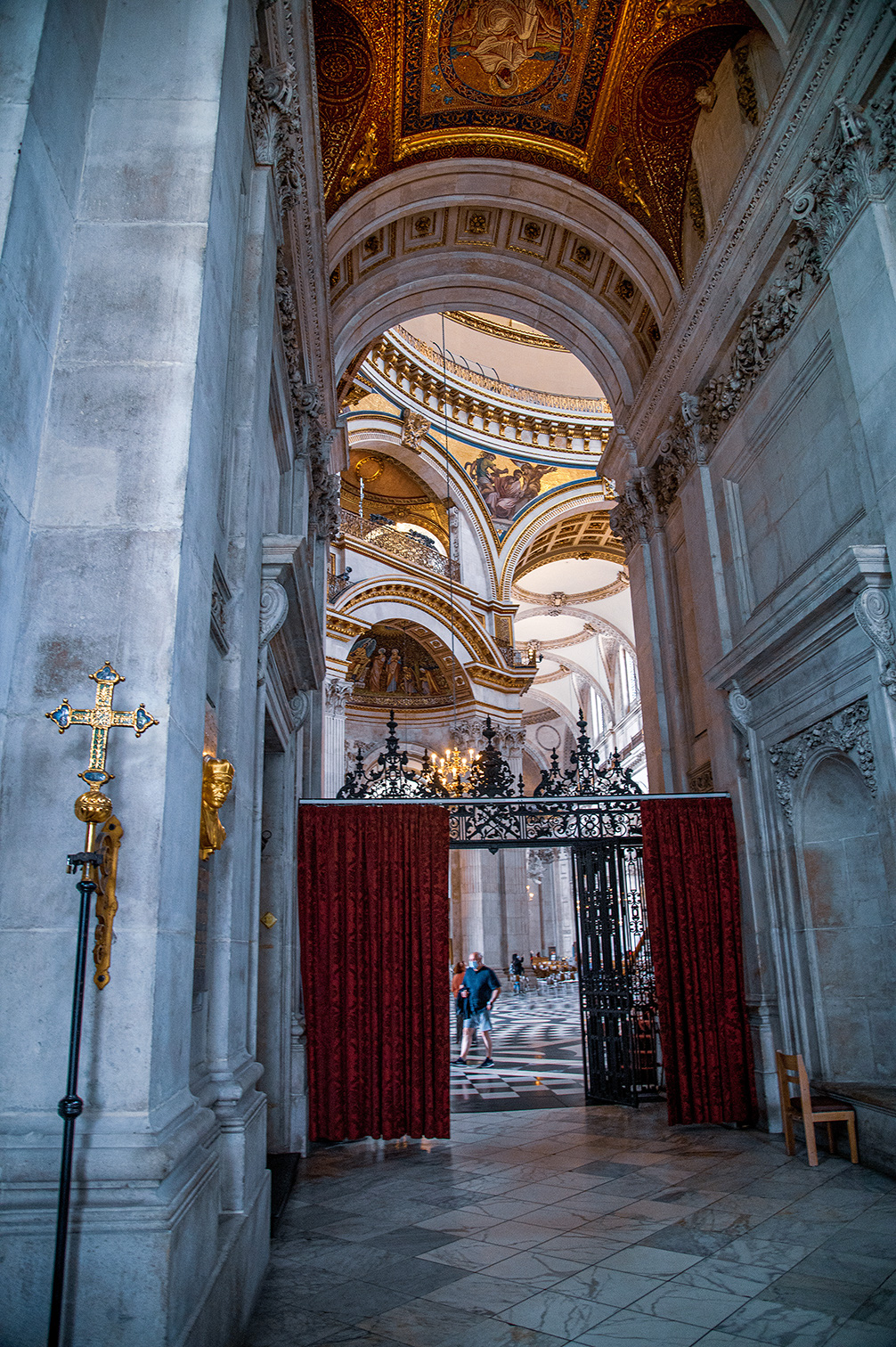The image depicts an ornate, grand interior of a historical building, likely a cathedral or church. The architecture features intricate stone carvings, arches, and a highly decorated ceiling with gold accents and detailed frescoes. The floor is tiled with a geometric pattern, and there is a large, ornate gate with red drapes. A person is seen walking through the distant end of the hall, which is well-lit by natural light coming through the windows.